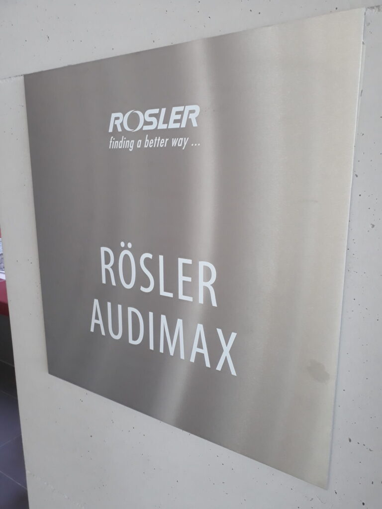 A metallic sign on a wall reads "Rösler Audimax" with the Rösler logo and slogan "finding a better way..." displayed above the text, showcasing their dedication to innovation in service centers. at Hochschule Coburg