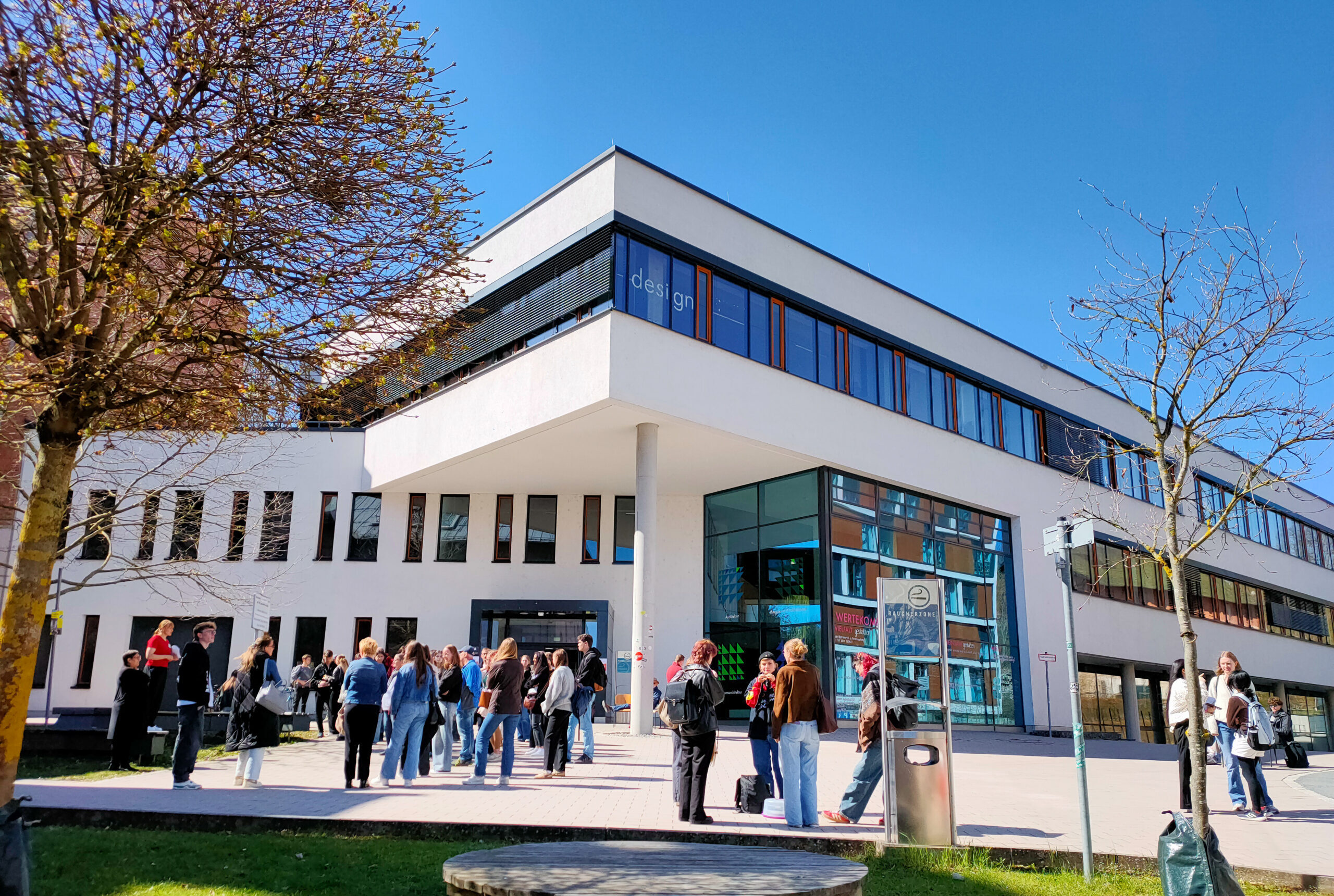 Eine Gruppe von Menschen erkundet die Campusluft vor einem modernen weißen Gebäude mit großen Fenstern unter einem klaren blauen Himmel. Einige Bäume säumen den gepflasterten Bereich, und das Sonnenlicht wirft Schatten auf den Boden. at Hochschule Coburg