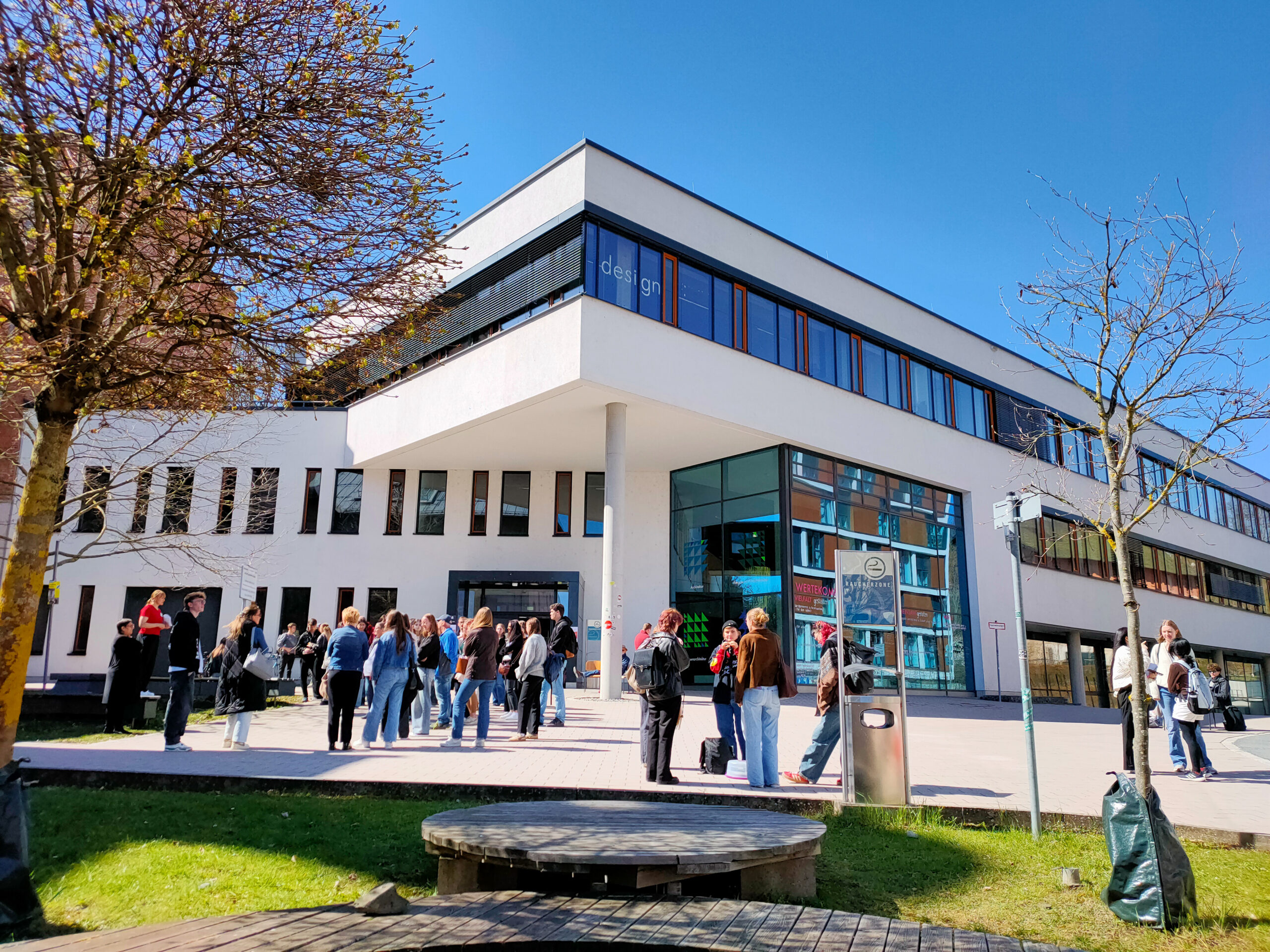 Eine Gruppe von Menschen erkundet die Campusluft vor einem modernen weißen Gebäude mit großen Fenstern unter einem klaren blauen Himmel. Einige Bäume säumen den gepflasterten Bereich, und das Sonnenlicht wirft Schatten auf den Boden. at Hochschule Coburg