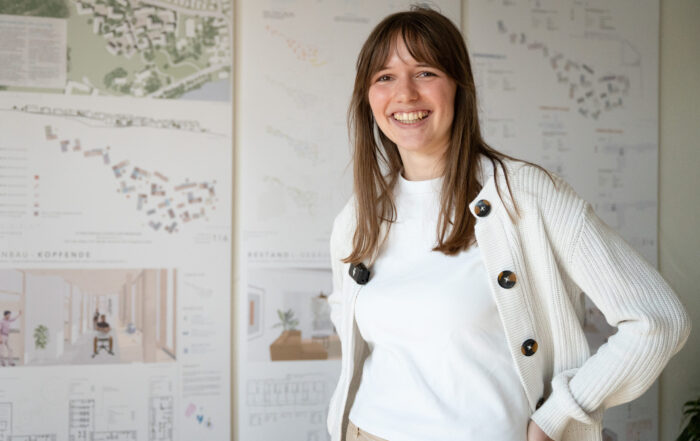 Eine junge Frau mit langen braunen Haaren steht lächelnd vor großen Plakaten, auf denen Architekturprojekt-Diagramme abgebildet sind. Sie trägt ein weißes Hemd und eine helle Strickjacke und steht mit einer Hand in der Hüfte. at Hochschule Coburg