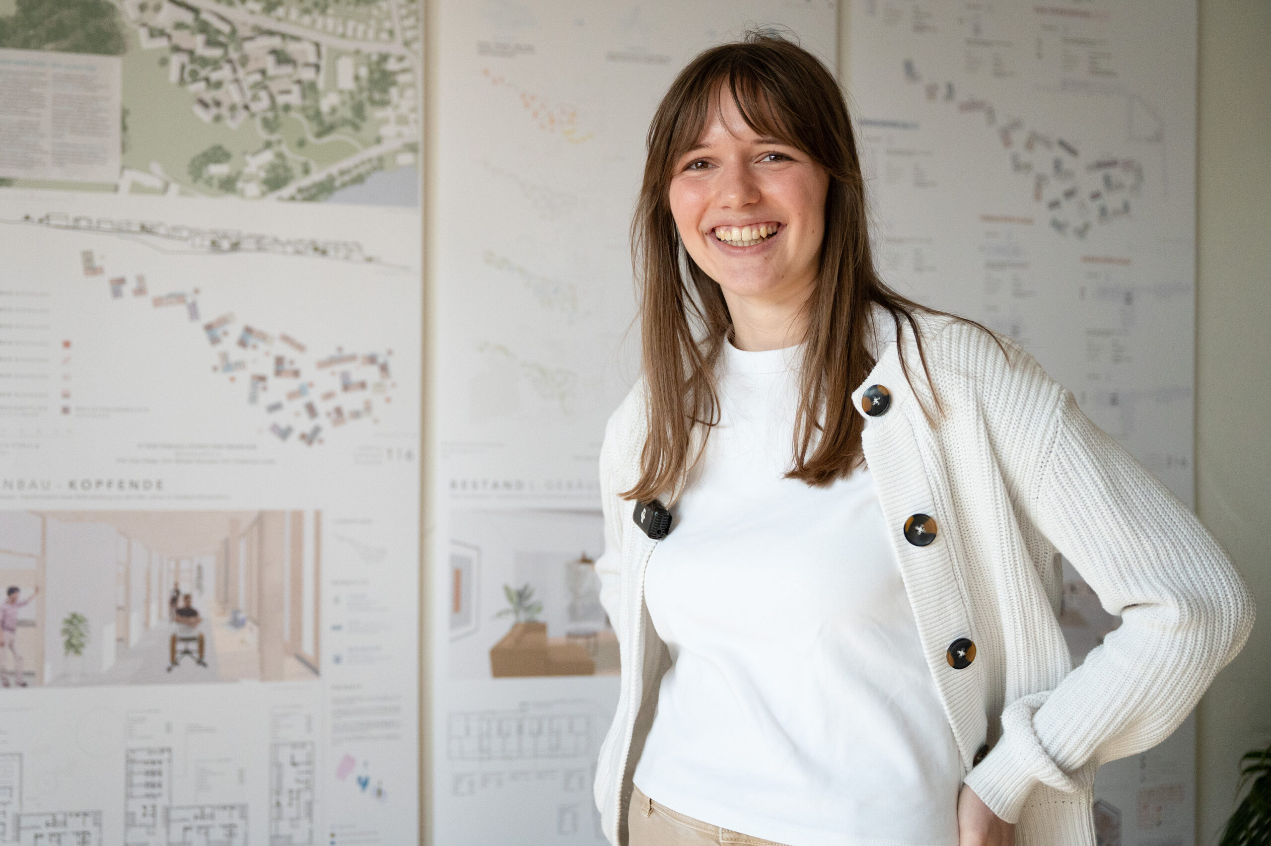 Eine junge Frau mit langen braunen Haaren steht lächelnd vor großen Plakaten, auf denen Architekturprojekt-Diagramme abgebildet sind. Sie trägt ein weißes Hemd und eine helle Strickjacke und steht mit einer Hand in der Hüfte. at Hochschule Coburg