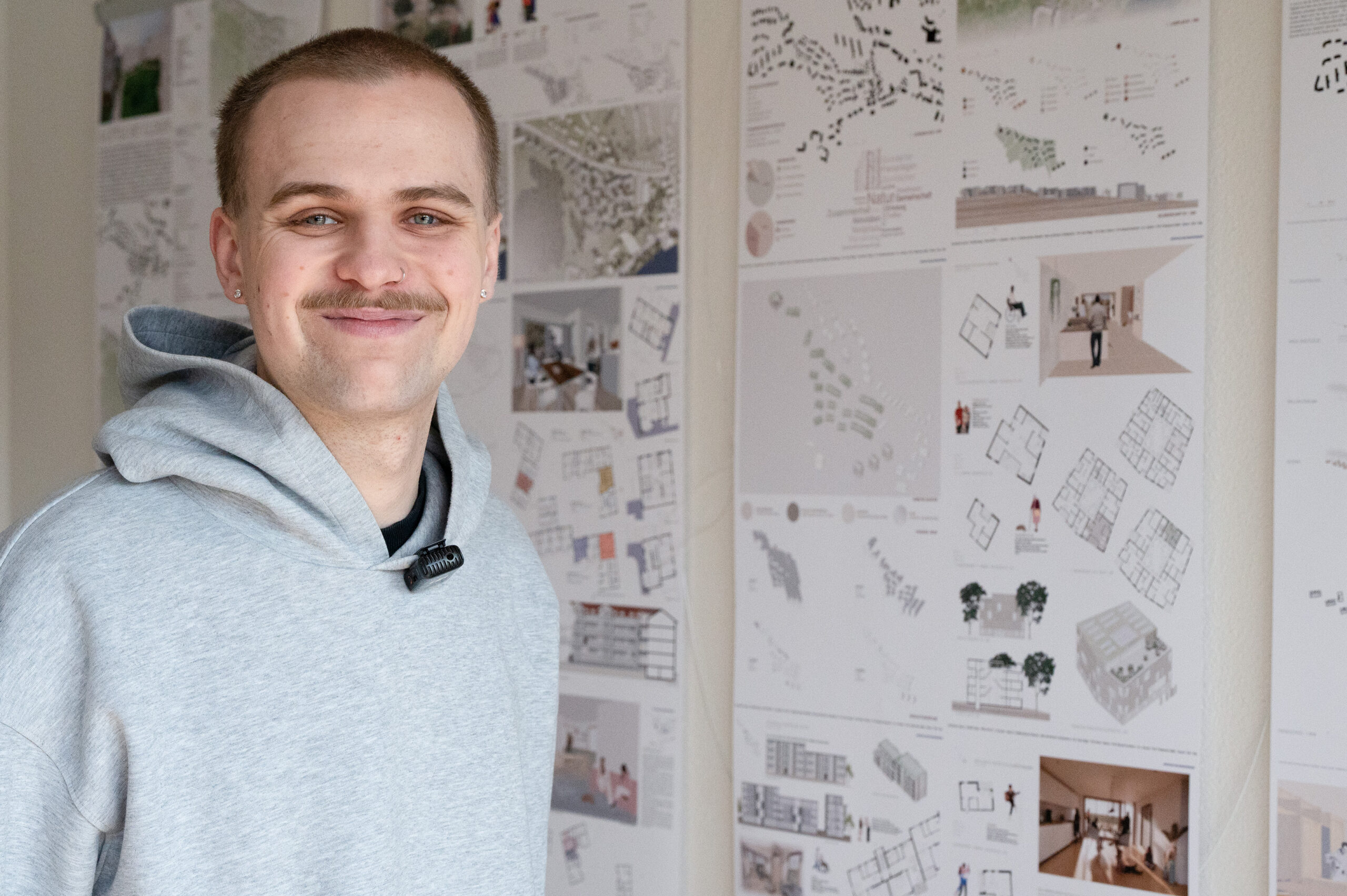 Eine Person in einem hellgrauen Kapuzenpulli lächelt in die Kamera. Sie steht vor einer Wand, die mit detaillierten Zeichnungen und Plänen des Architekturprojekts bedeckt ist. at Hochschule Coburg