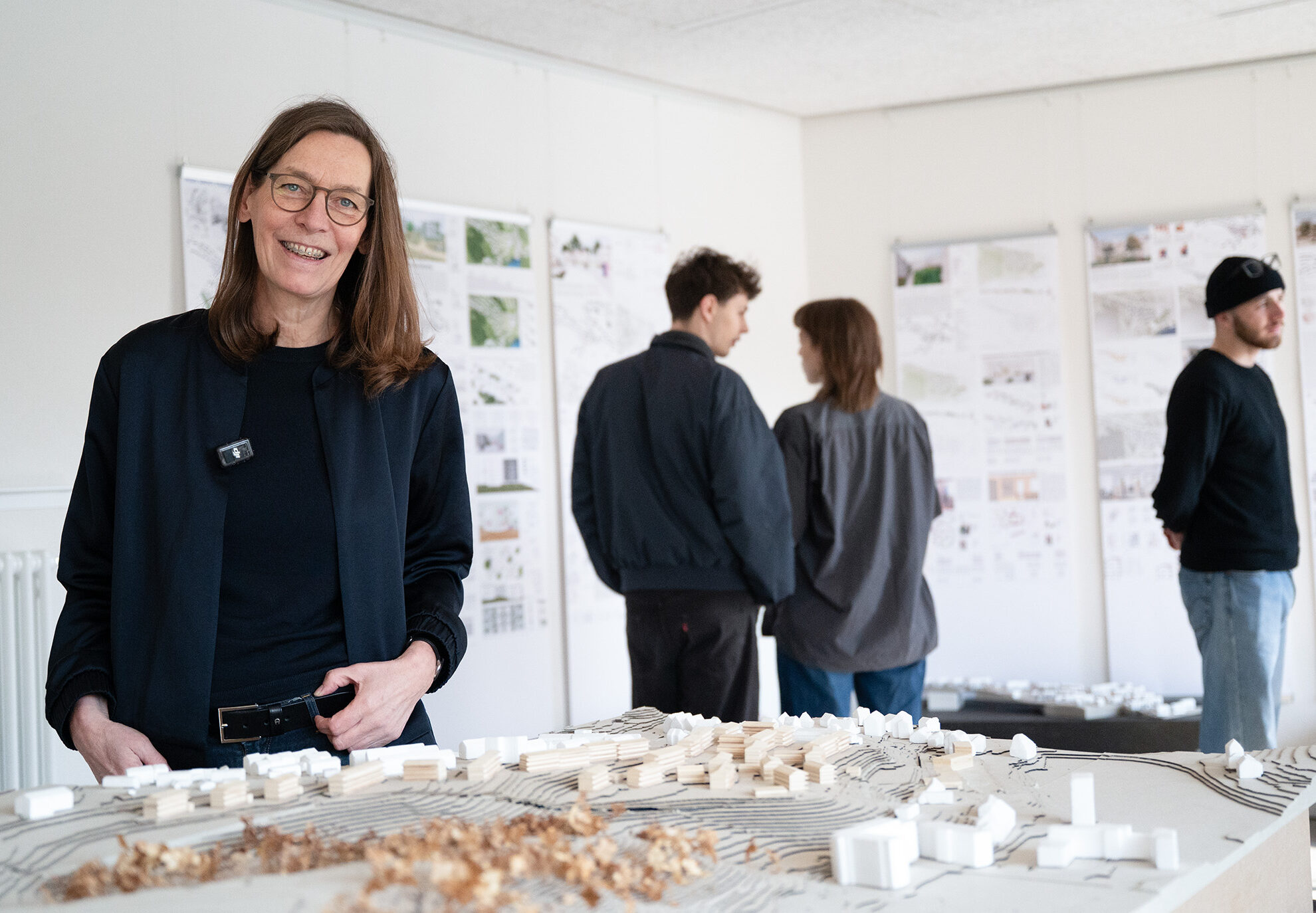 Eine Frau steht lächelnd neben einem Architekturmodell auf einem Tisch, das Teil des Architekturprojekts in Wüstenahorn ist, während drei Personen im Hintergrund Zeichnungen und Pläne betrachten, die an den Wänden eines hellen Raums angebracht sind. at Hochschule Coburg