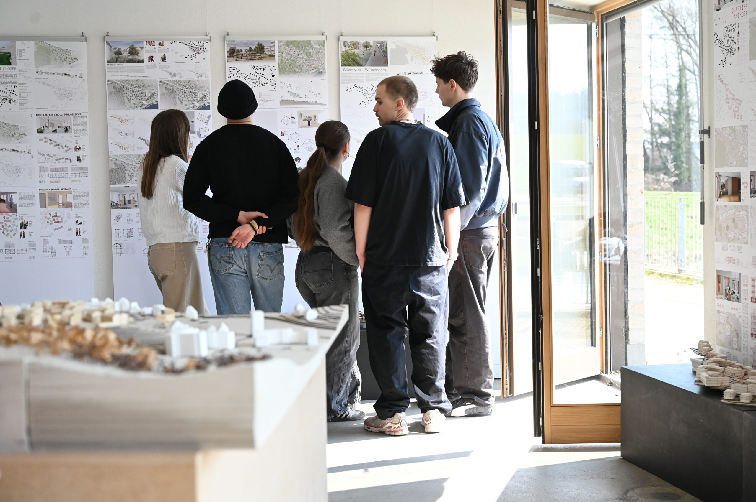 Sechs Personen stehen in einem hellen Raum und betrachten die Zeichnungen und Pläne des Architekturprojekts, die an der Wand hängen; im Vordergrund ist ein großes Architekturmodell von Wüstenahorn zu sehen. at Hochschule Coburg