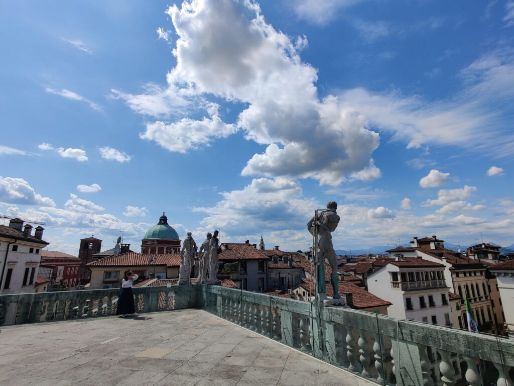 Eine Terrasse mit Statuen blickt auf eine Stadtlandschaft mit roten Dächern unter einem strahlend blauen Himmel mit vereinzelten Wolken. Eine Person steht in der Nähe der Statuen und blickt auf die Aussicht. at Hochschule Coburg
