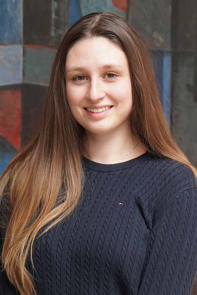 Eine junge Frau mit langen braunen Haaren und einem dunkelblauen Pullover lächelt in die Kamera. Sie steht vor einem farbenfrohen abstrakten Wandgemälde, das ihre Leidenschaft für Informatik und Naturwissenschaften widerspiegelt. at Hochschule Coburg