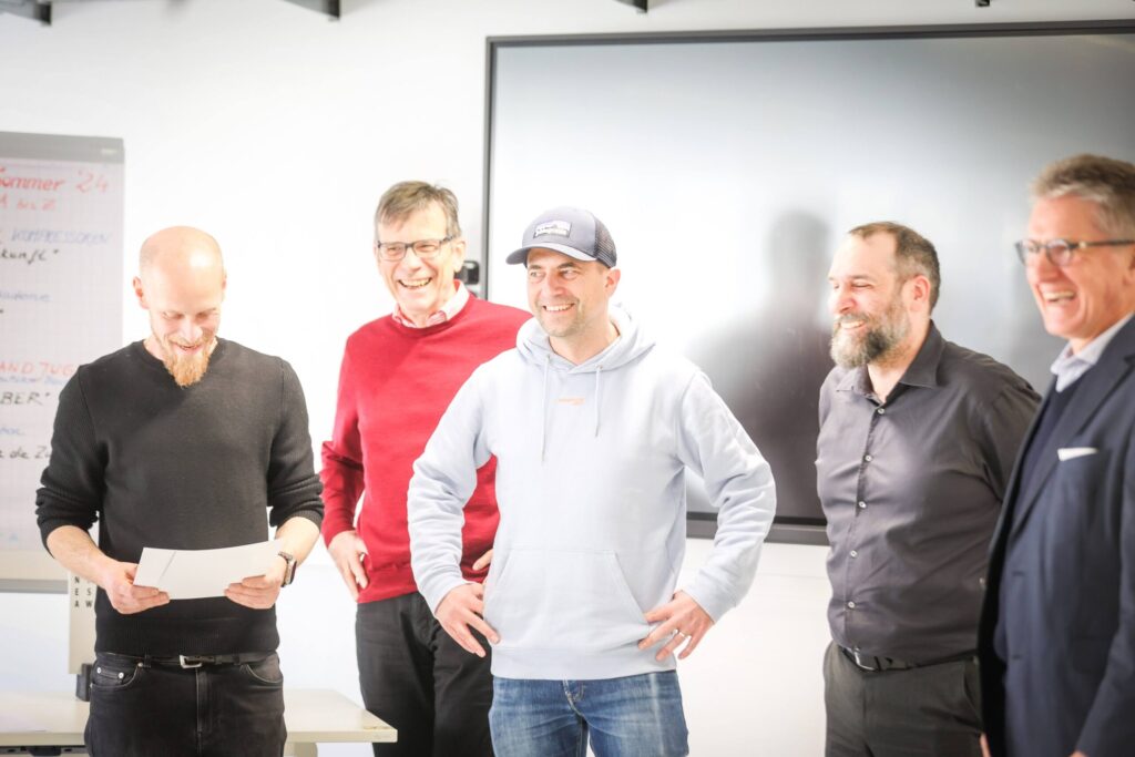 Fünf Männer stehen in einem Innenraum zusammen, lächeln und lachen. Ein Mann auf der linken Seite hält ein Blatt Papier in der Hand und blickt darauf hinunter, während die anderen zu ihm oder in die Kamera schauen, was den Geist der Innovation in einem Büro oder Besprechungsraum widerspiegelt. at Hochschule Coburg
