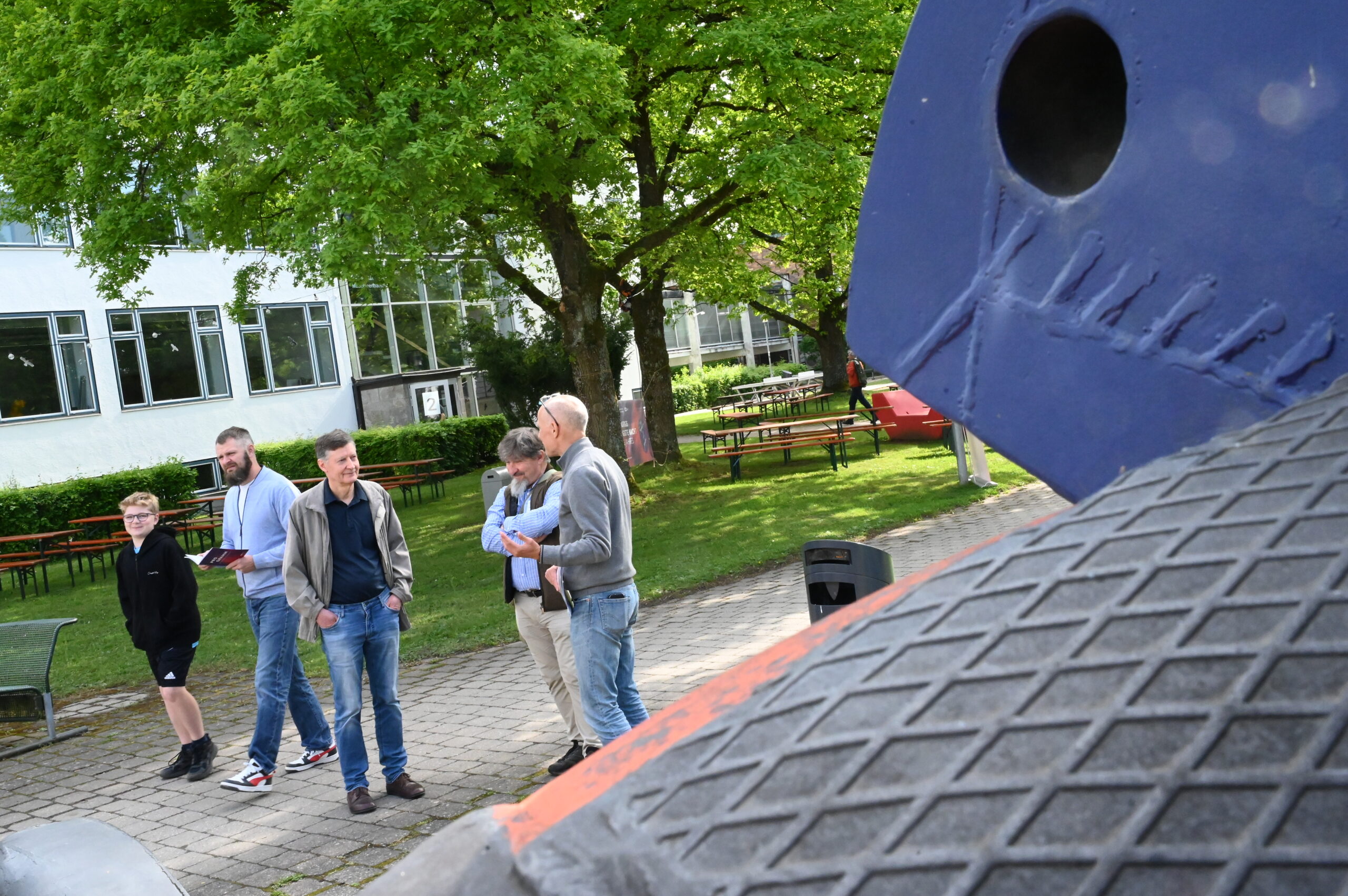 Eine Gruppe von fünf Personen steht während der Langen Nacht der Wissenschaften draußen in der Nähe einer großen Skulptur und unterhält sich. Im Hintergrund stehen Bäume und ein Gebäude mit großen Fenstern, und auf dem Rasen sind Bänke zu sehen, die eine Atmosphäre wissenschaftlicher Resonanz schaffen. at Hochschule Coburg