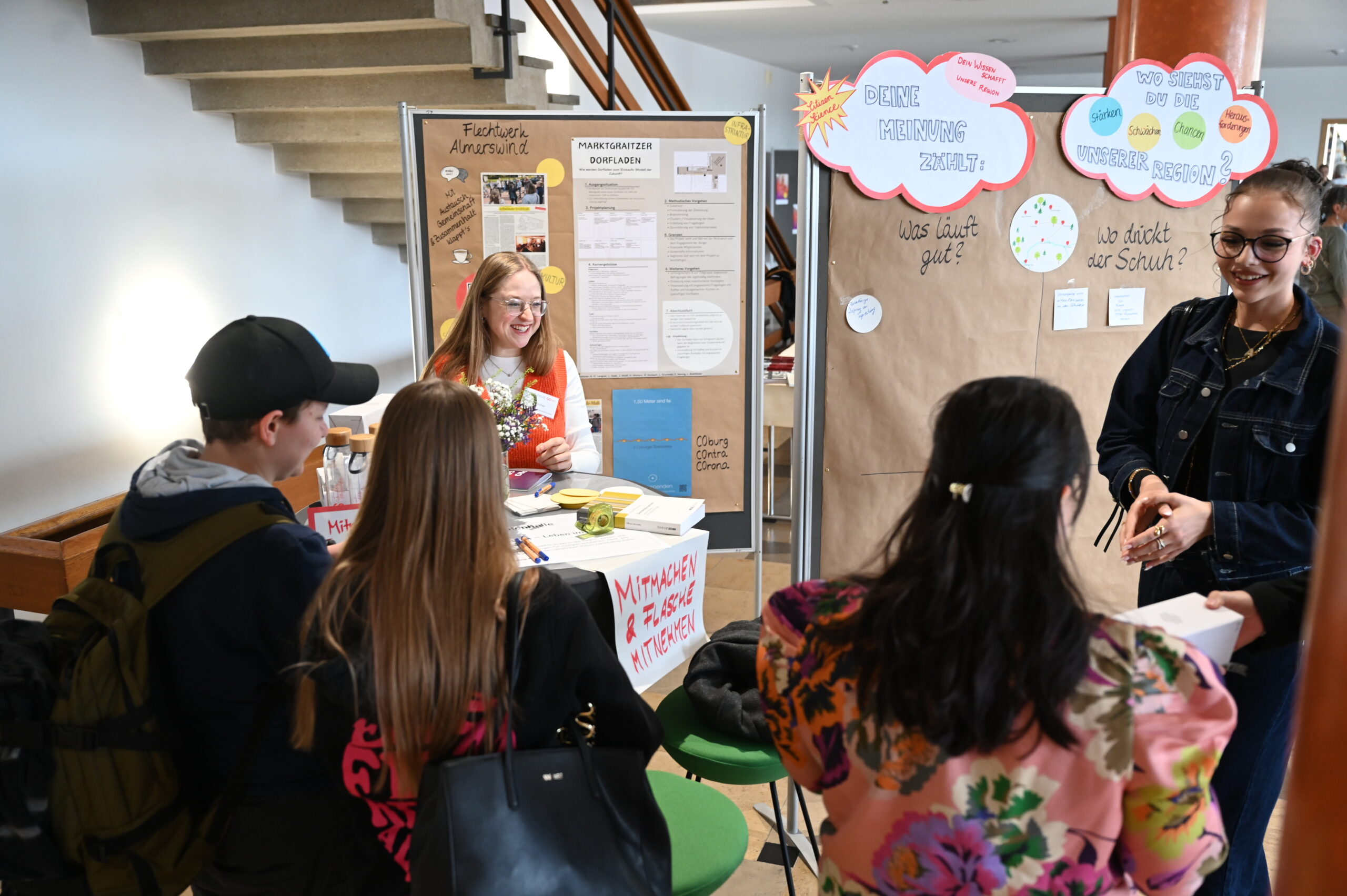 Vier Personen stehen um einen Stand mit Plakaten und Informationstafeln und unterhalten sich mit einer lächelnden Frau am Tisch. Der überdachte Stand in der Nähe einer Treppe zeigt Lehrmaterial für die Lange Nacht der Wissenschaften und fördert so die Resonanz der Besucher. at Hochschule Coburg
