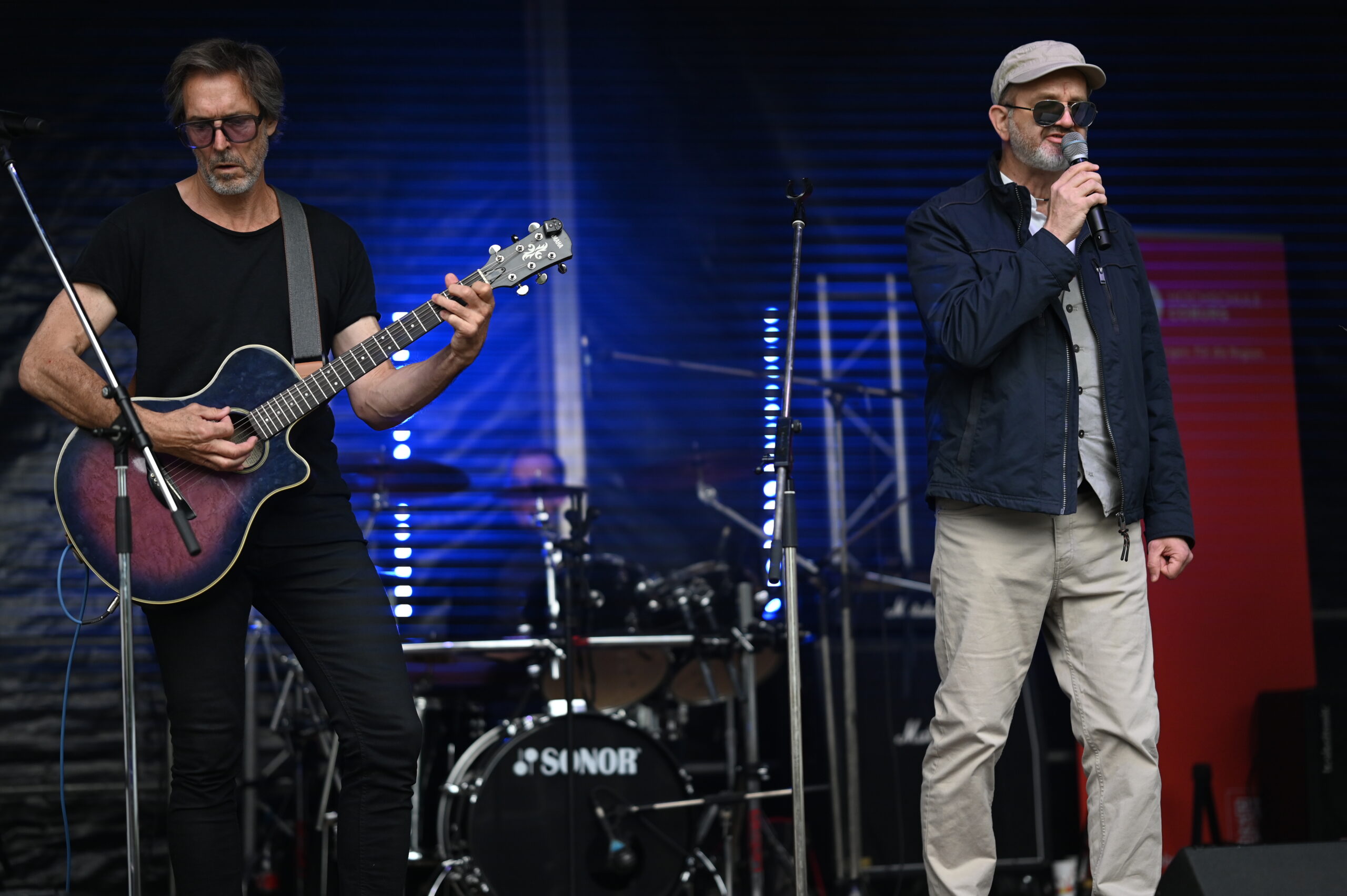Zwei Männer treten bei der Langen Nacht der Wissenschaften auf; der eine spielt in Sonnenbrille und schwarzem Hemd eine Akustikgitarre, der andere singt mit Mütze und Jacke in ein Mikrofon. Ein Schlagzeug trägt zur Resonanz im Hintergrund bei. at Hochschule Coburg
