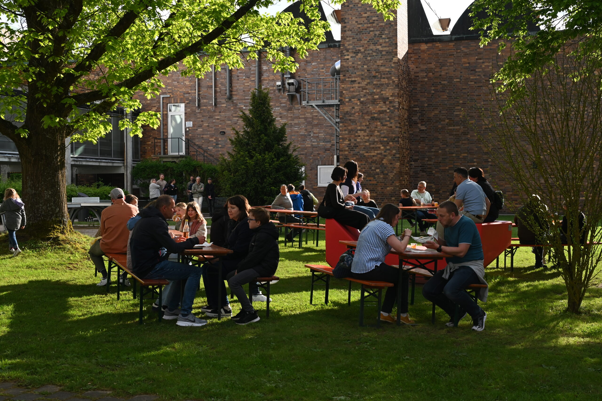 Während der Langen Nacht der Wissenschaften sitzen Menschen an roten Picknicktischen auf einer Wiese und unterhalten sich bei Getränken im Schatten eines großen Baumes, während sich an diesem sonnigen Tag weitere Menschen in der Nähe eines Backsteingebäudes im Hintergrund versammeln. at Hochschule Coburg