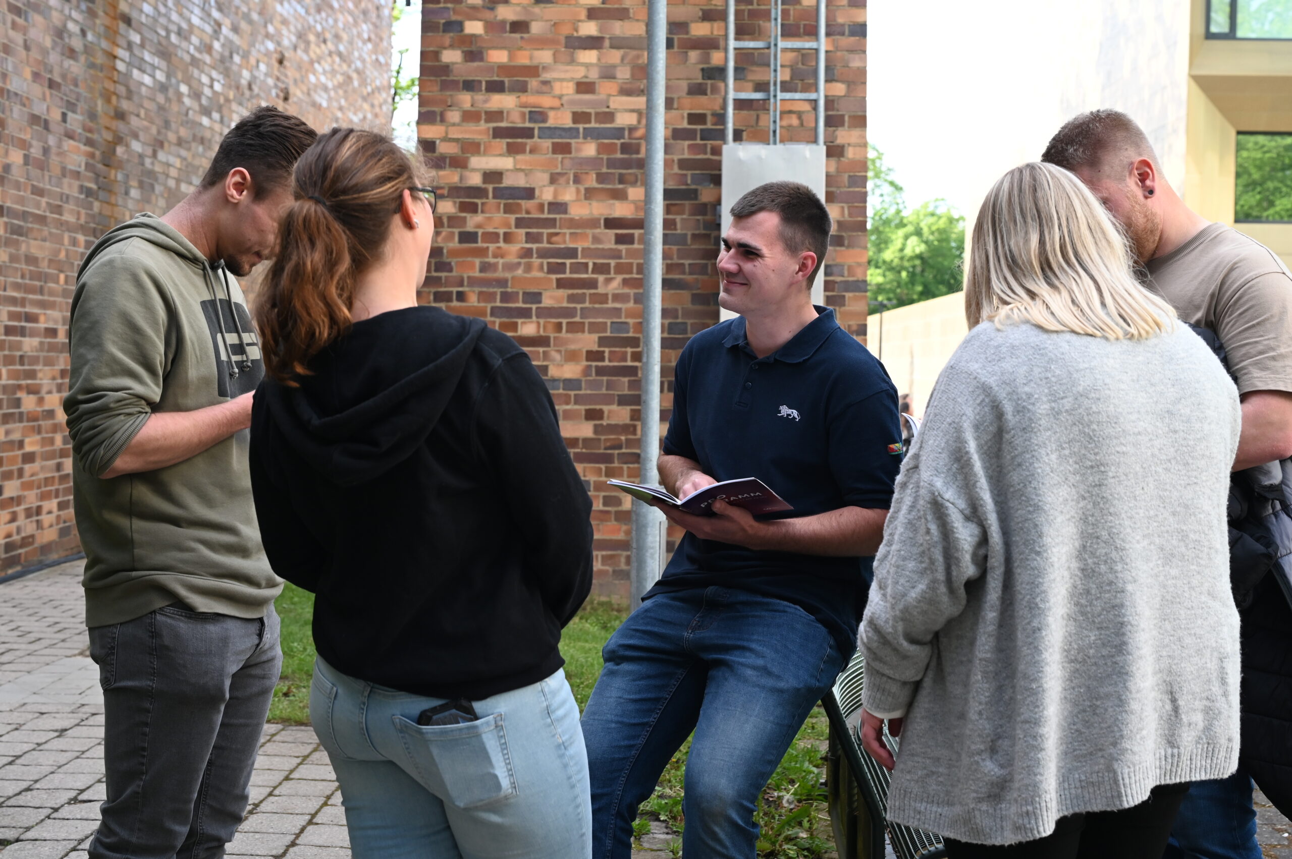 Eine Gruppe von fünf Erwachsenen steht während der Langen Nacht der Wissenschaften draußen in der Nähe von Backsteingebäuden und unterhält sich; eine Person sitzt auf einer niedrigen Mauer und hält ein aufgeschlagenes Notizbuch in der Hand, während die anderen zuhören und so die Resonanz der Neugierigen fördern. at Hochschule Coburg