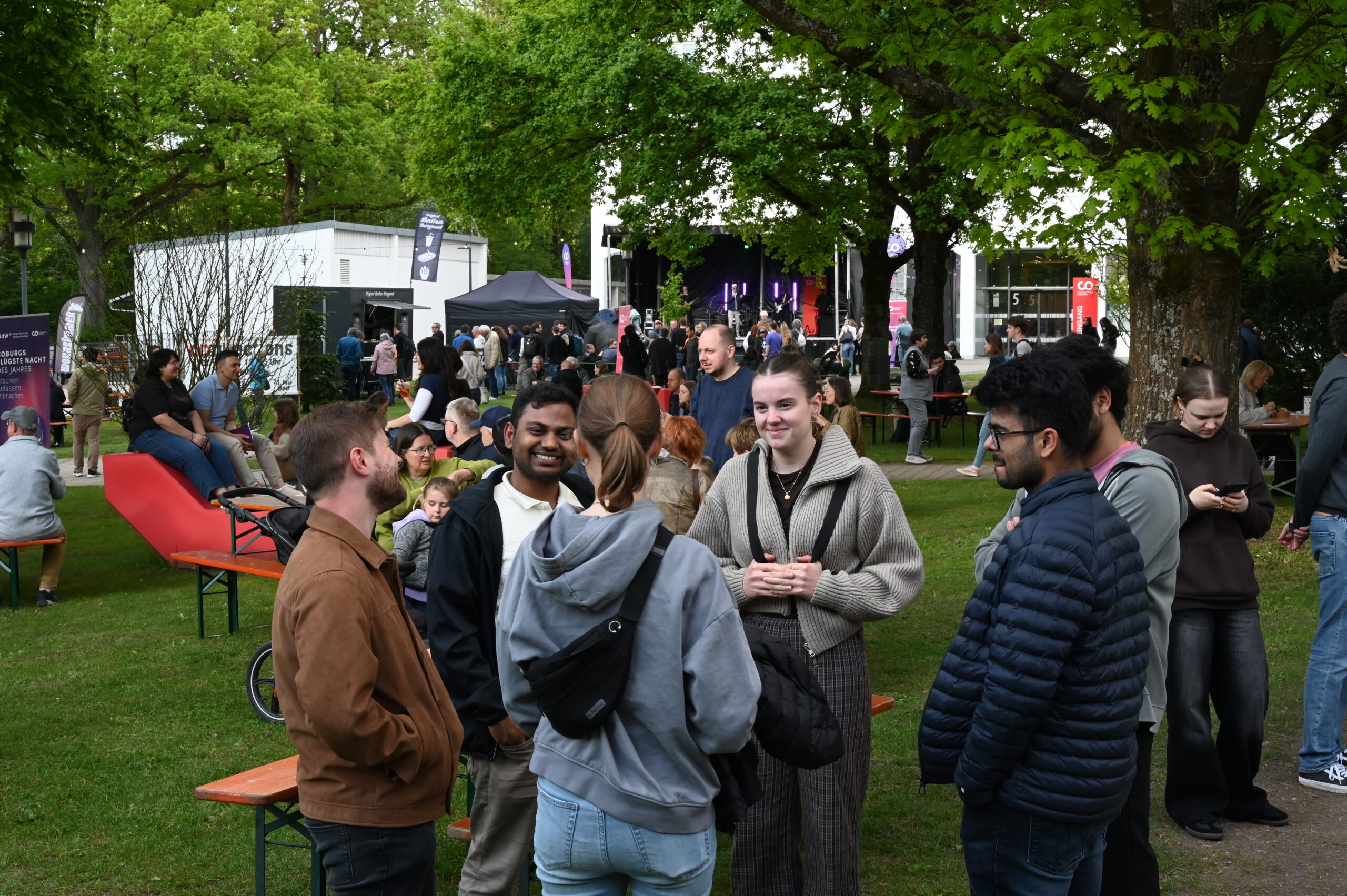 Eine Gruppe von Menschen steht und unterhält sich auf einer Wiese bei der Langen Nacht der Wissenschaften, mit Bäumen, Bänken und Menschenmengen im Hintergrund. Einige lächeln; weiter hinten sind Zelte, eine Bühne und die lebhafte Resonanz der Wissenschaft zu sehen. at Hochschule Coburg
