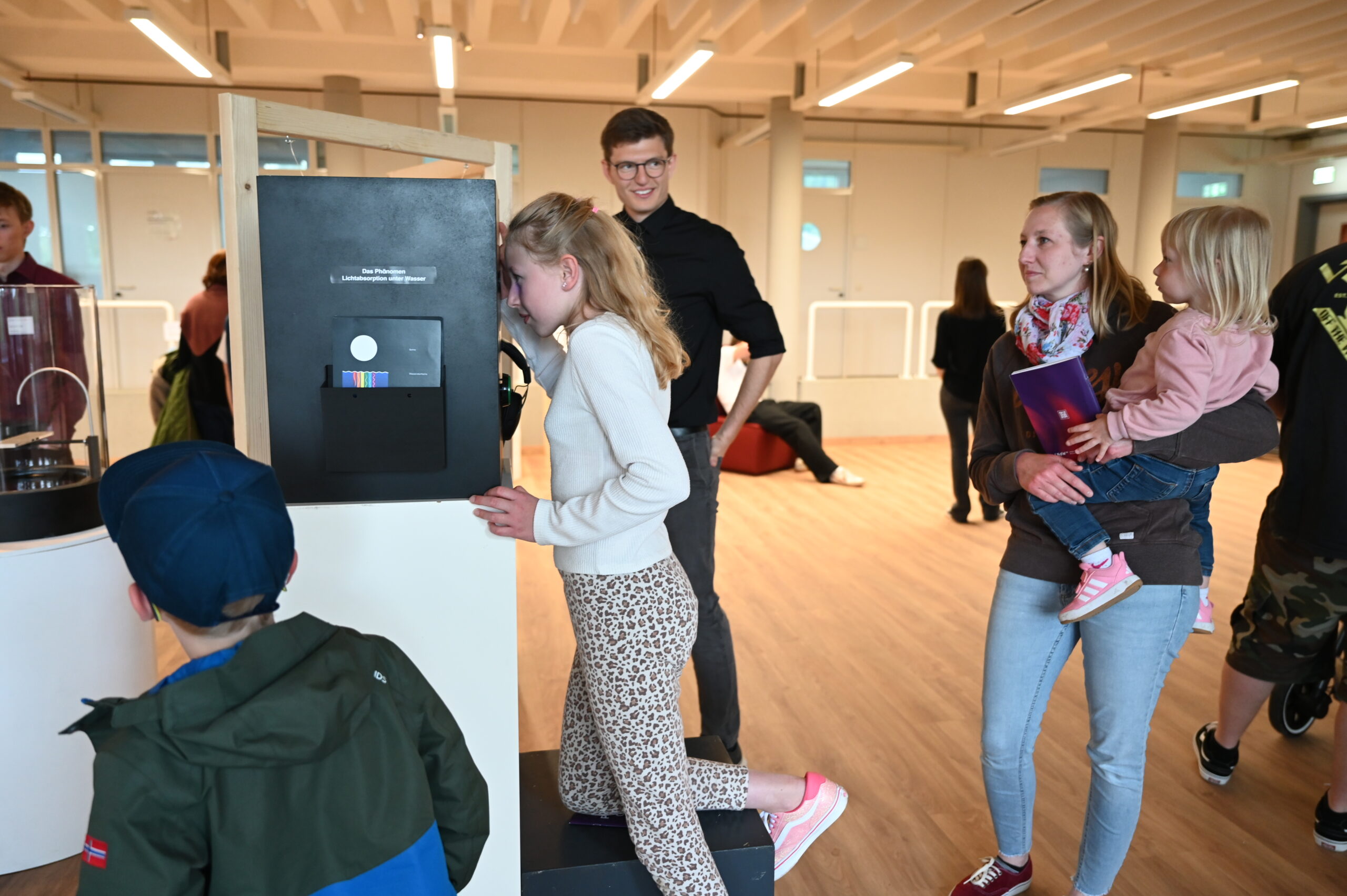 In der Langen Nacht der Wissenschaften blickt ein Mädchen in einen schwarzen Schaukasten in einem modernen Innenraum, während ein lächelnder Mann sie beobachtet. In der Nähe hält eine Frau ein kleines Kind und ein Junge mit einer blauen Mütze steht in der Nähe. Im Hintergrund sind weitere Personen zu sehen. at Hochschule Coburg