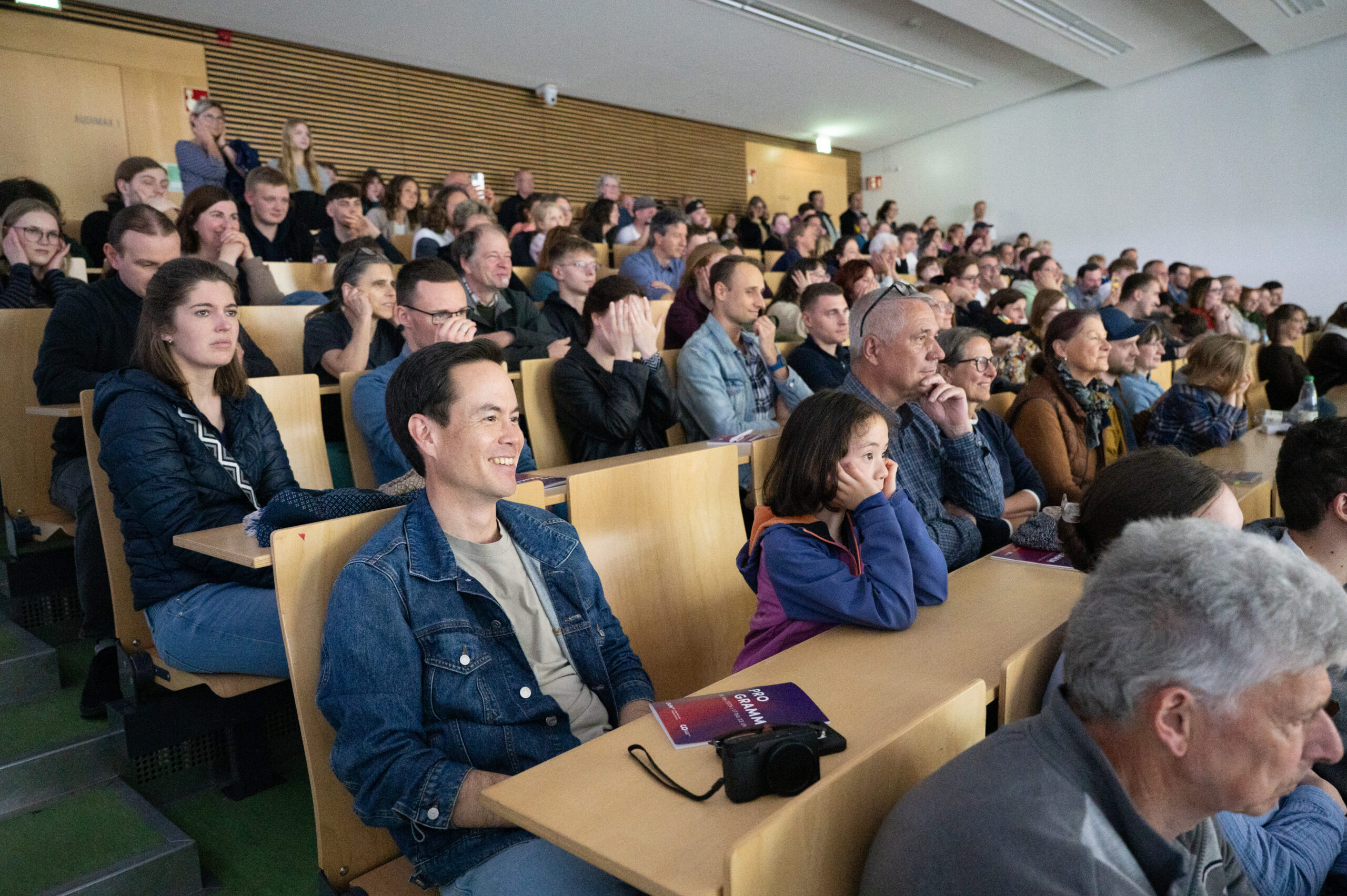 Eine große Gruppe von Menschen unterschiedlichen Alters sitzt während der Langen Nacht der Wissenschaften in gestaffelten Reihen in einem Hörsaal und schaut aufmerksam nach vorne. Einige klatschen oder jubeln, andere wirken konzentriert und erzeugen eine lebhafte Resonanz im Raum. at Hochschule Coburg