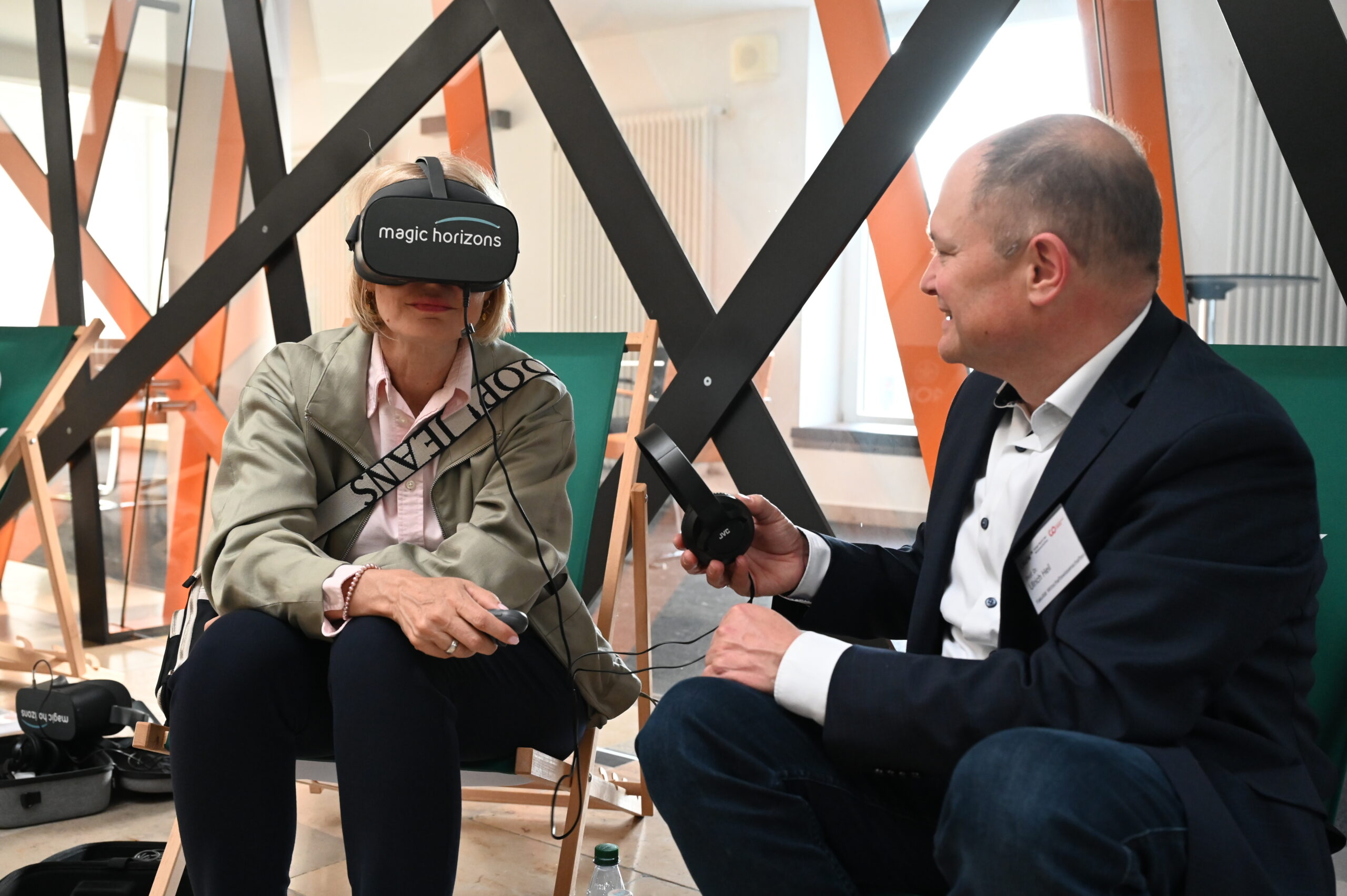 Eine Frau, die ein Virtual-Reality-Headset mit der Aufschrift "magic horizons" trägt, sitzt auf einem Stuhl, während ein Mann im Anzug bei der Langen Nacht der Wissenschaften einen VR-Controller hält und mit ihr in einem modernen Raum mit geometrischem Dekor spricht. at Hochschule Coburg
