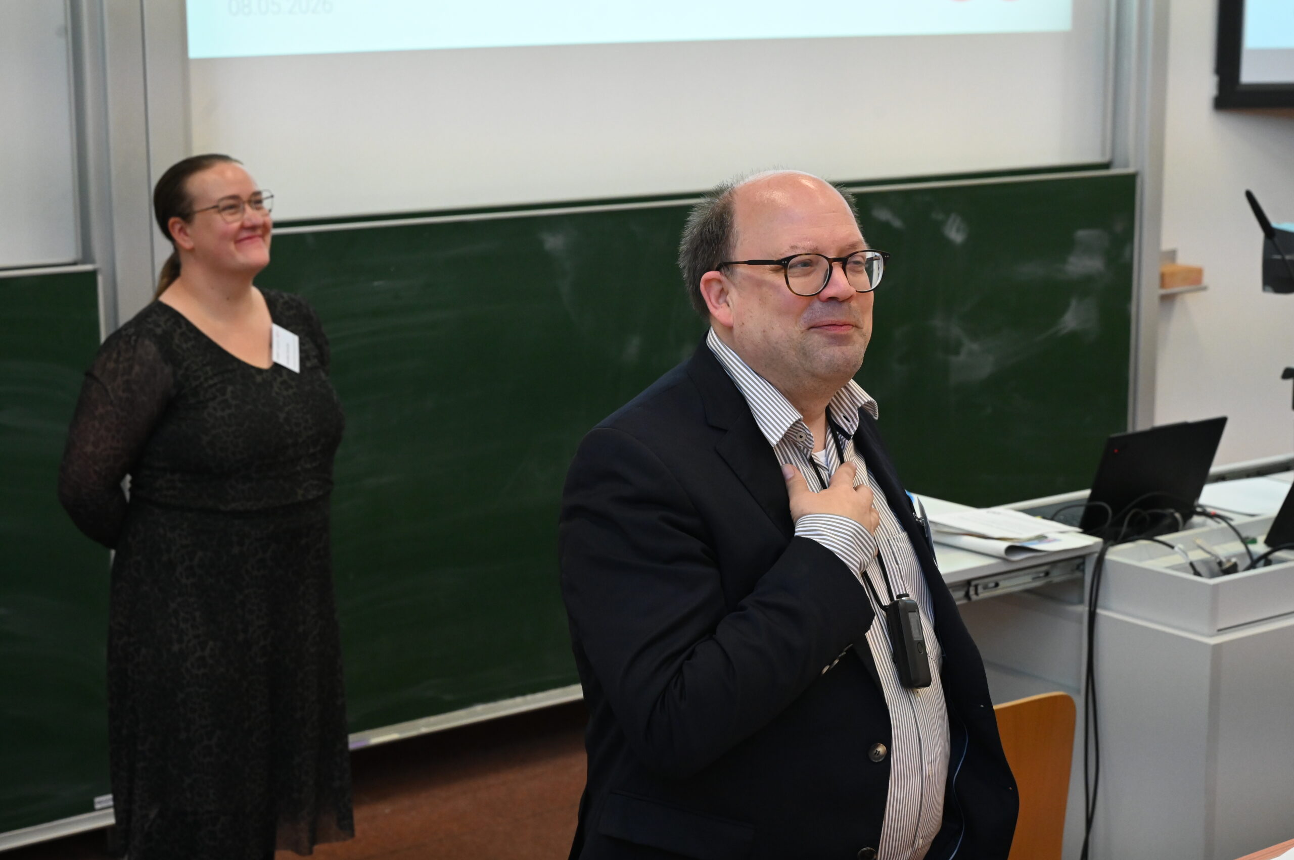 Ein Mann im Anzug steht vor einer Kreidetafel und spricht bei der Langen Nacht der Wissenschaften, wobei er ein Mikrofon am Revers trägt. Eine Frau in einem schwarzen Kleid steht lächelnd im Hintergrund neben einem Schreibtisch und einem Laptop. at Hochschule Coburg