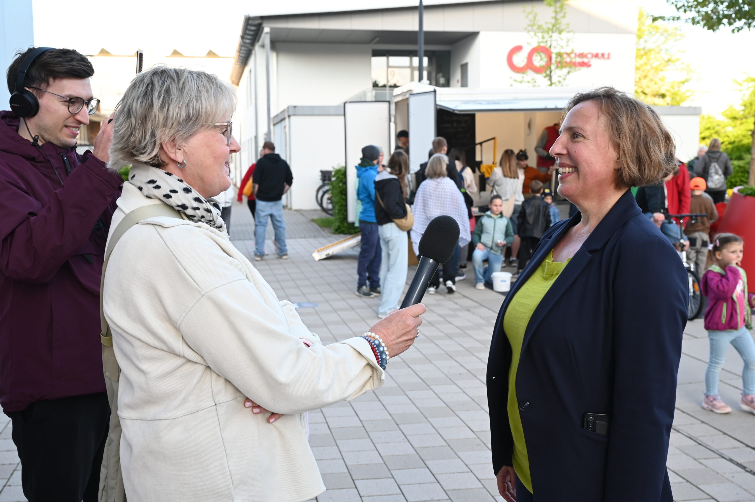 Eine Frau mit einem Mikrofon interviewt eine andere Frau im Freien vor einer Menschenmenge bei der Langen Nacht der Wissenschaften. Die Menschen haben sich in der Nähe eines modernen Gebäudes versammelt, und ein Mann mit Kopfhörern steht in der Nähe und nimmt das Interview möglicherweise für Resonanz auf. at Hochschule Coburg