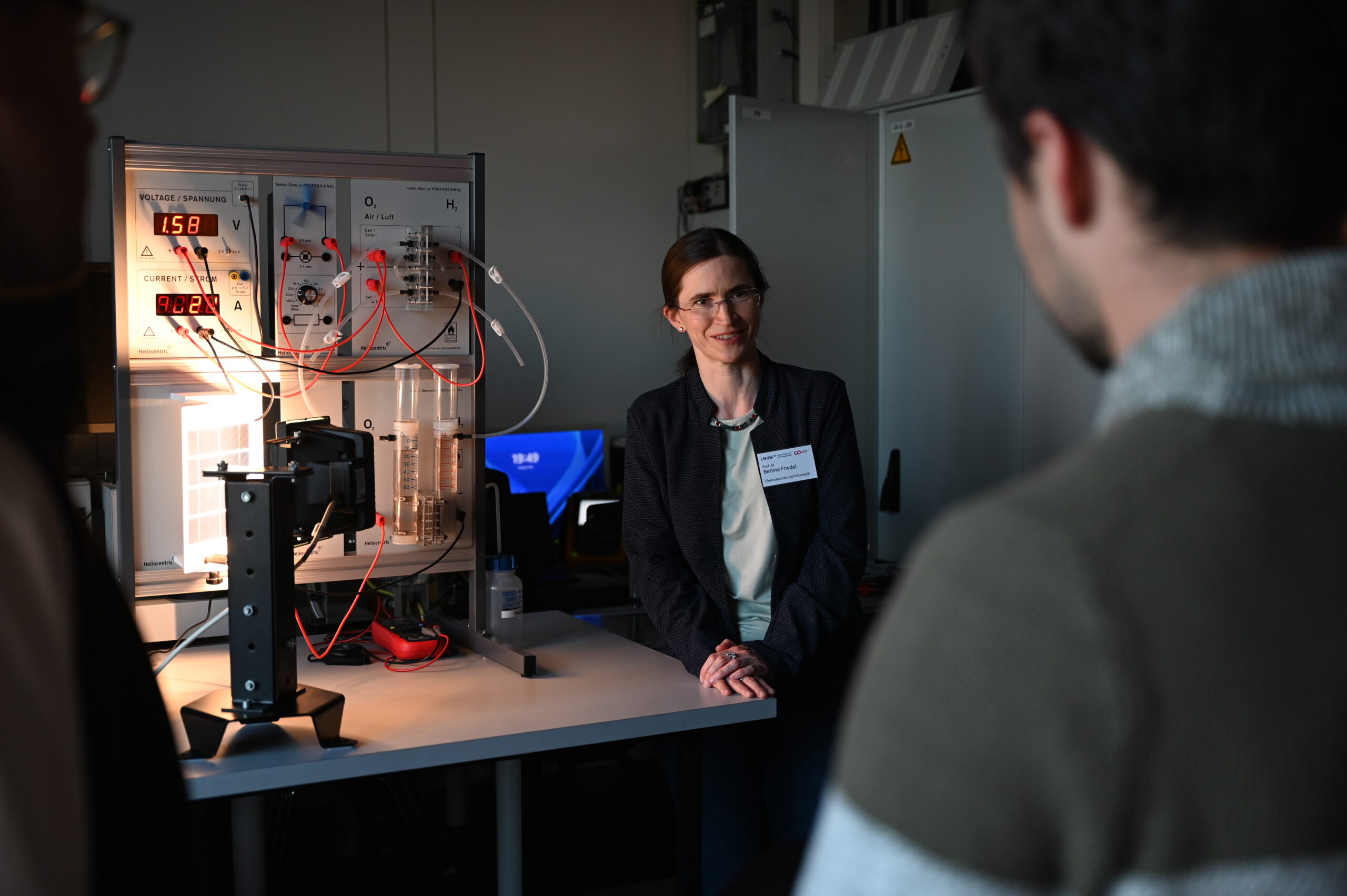 In der Langen Nacht der Wissenschaften steht eine Frau mit Brille und dunklem Blazer an einem Laborarbeitsplatz und diskutiert mit zwei Personen über Wissenschaft und Resonanz. In dem schwach beleuchteten Labor sind im Hintergrund wissenschaftliche Geräte, Leitungen und Kabel zu sehen. at Hochschule Coburg