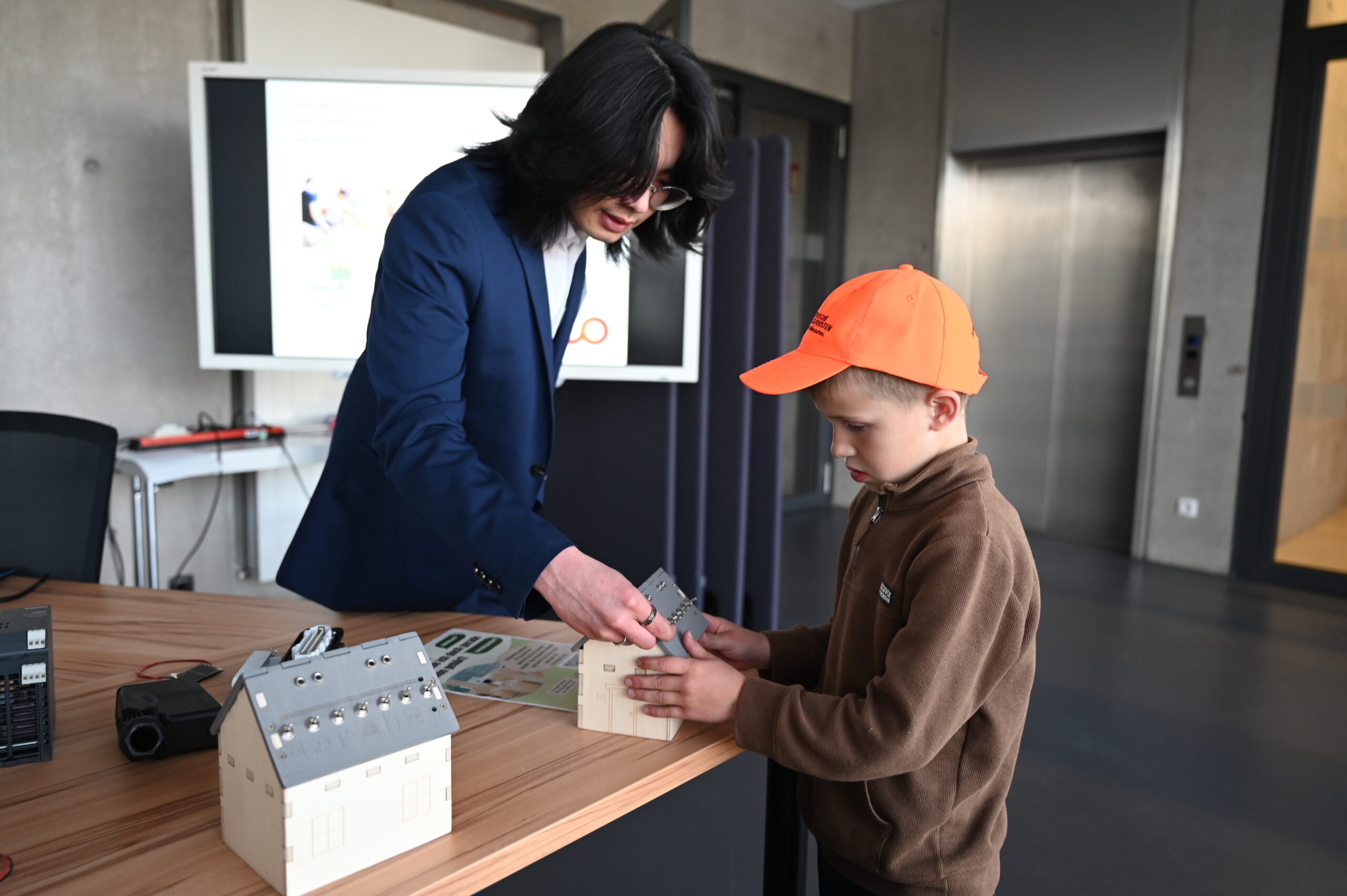 Bei der Langen Nacht der Wissenschaften hilft ein Erwachsener in einem blauen Blazer einem Jungen mit einer orangefarbenen Mütze, ein Modellhaus auf einem Tisch zusammenzubauen. In einem modernen Raum mit einem weiteren Modellhaus und einem Präsentationsbildschirm im Hintergrund schwingen Neugierde und Lernbereitschaft mit. at Hochschule Coburg