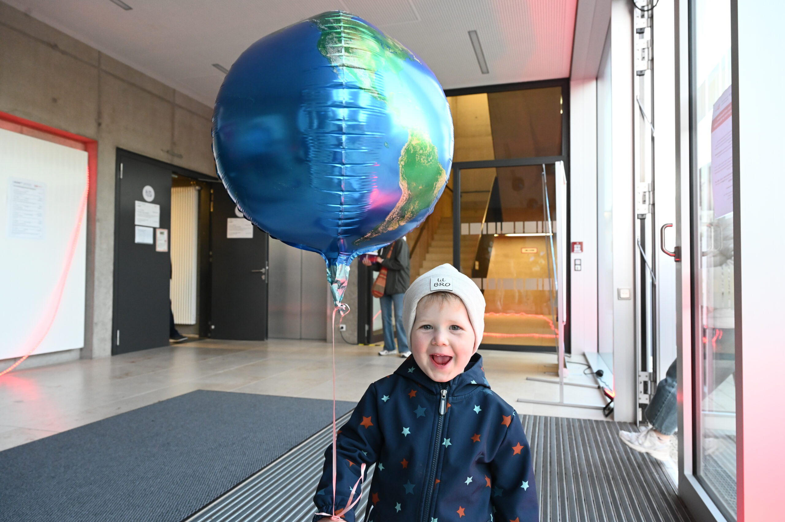 Ein lächelndes Kind mit Sternenmantel und weißem Hut hält einen großen Luftballon in der Hand. Es steht im Inneren eines modernen Gebäudes in der Nähe von Glastüren während der Langen Nacht der Wissenschaften, die die Freude an der Wissenschaft für alle Altersgruppen feiert. at Hochschule Coburg