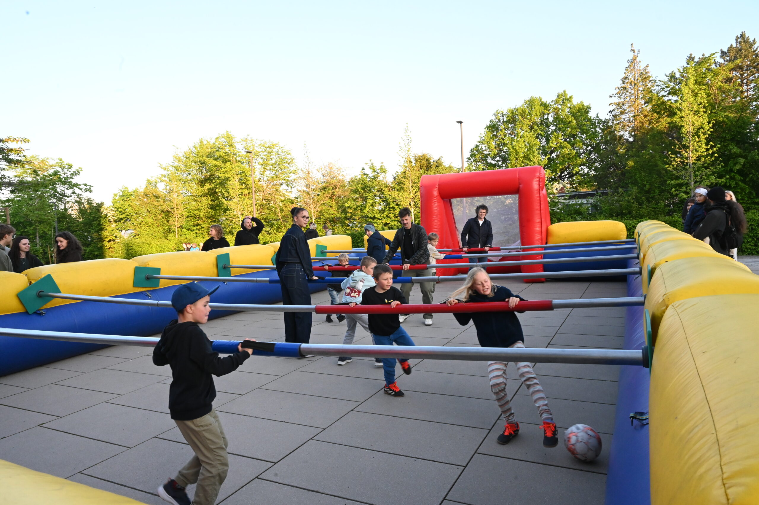 Kinder spielen während der Langen Nacht der Wissenschaften im Freien ein lebensgroßes aufblasbares menschliches Tischfußballspiel, wobei sich jeder an horizontalen Stangen festhält. Erwachsene und andere Kinder schauen zu und tragen zur lebendigen Resonanz unter Bäumen und blauem Himmel bei. at Hochschule Coburg
