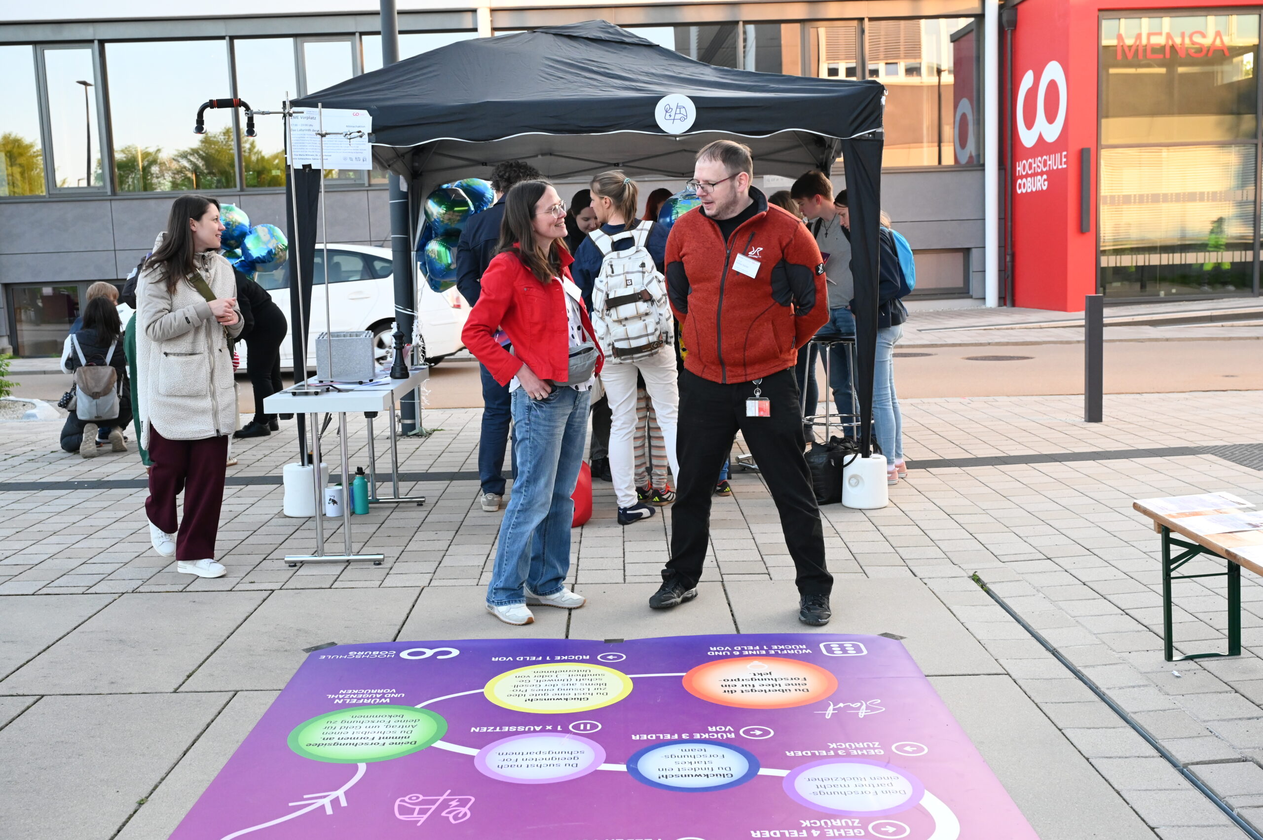 Menschen stehen und unterhalten sich bei einer Veranstaltung der Langen Nacht der Wissenschaften im Freien in der Nähe eines Standes mit Luftballons und Informationsmaterial. Eine große bunte Matte mit Symbolen und Text liegt auf dem Boden vor dem modernen "Mensa"-Gebäude. at Hochschule Coburg