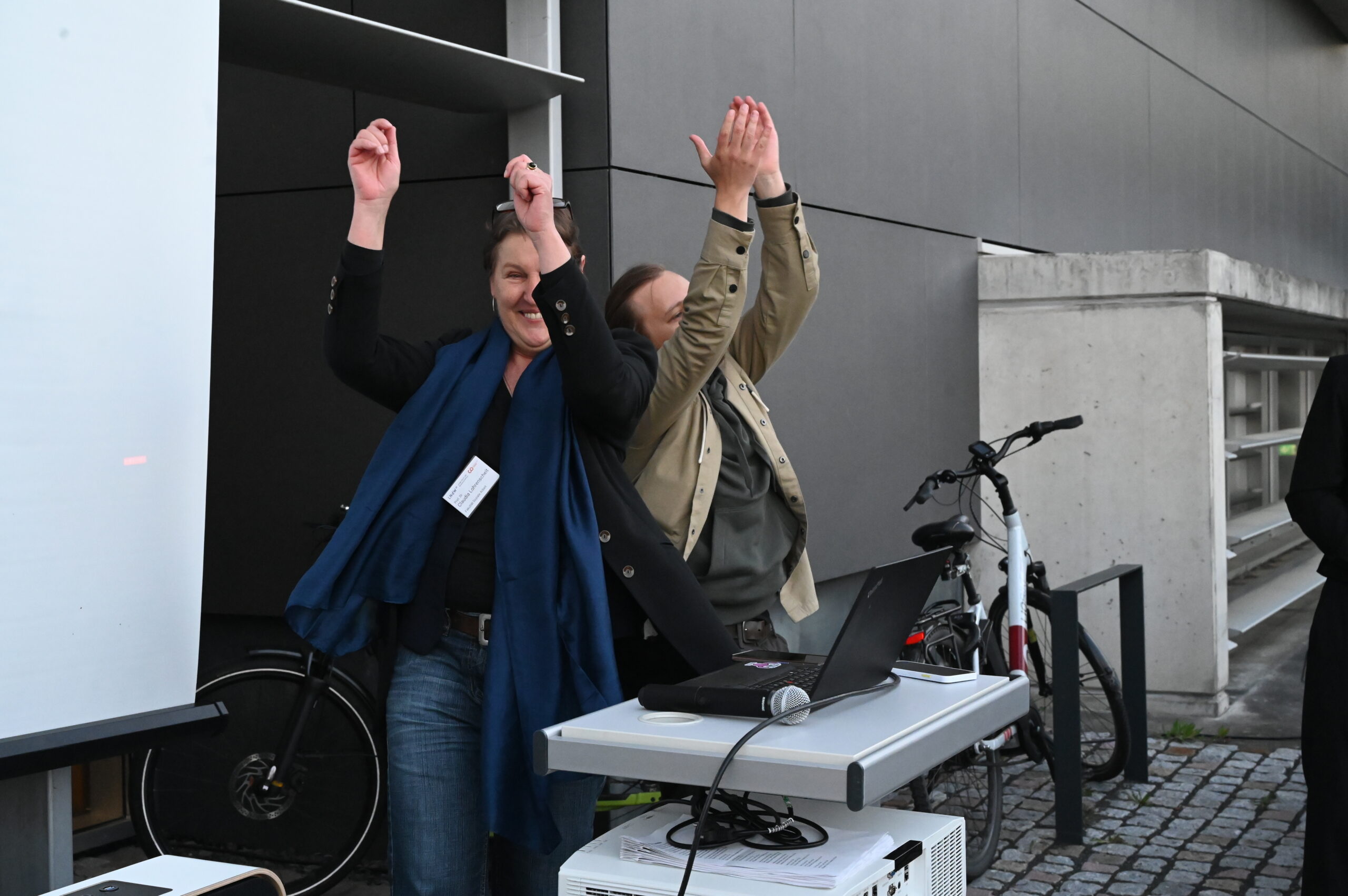 Zwei Menschen stehen im Freien nebeneinander, lächeln und heben feierlich die Arme in der Nähe eines Tisches mit einem Laptop und einem Projektor - sie fangen den Geist der Resonanz bei der Langen Nacht der Wissenschaften in einer städtischen Umgebung mit Beton und moderner Architektur ein. at Hochschule Coburg
