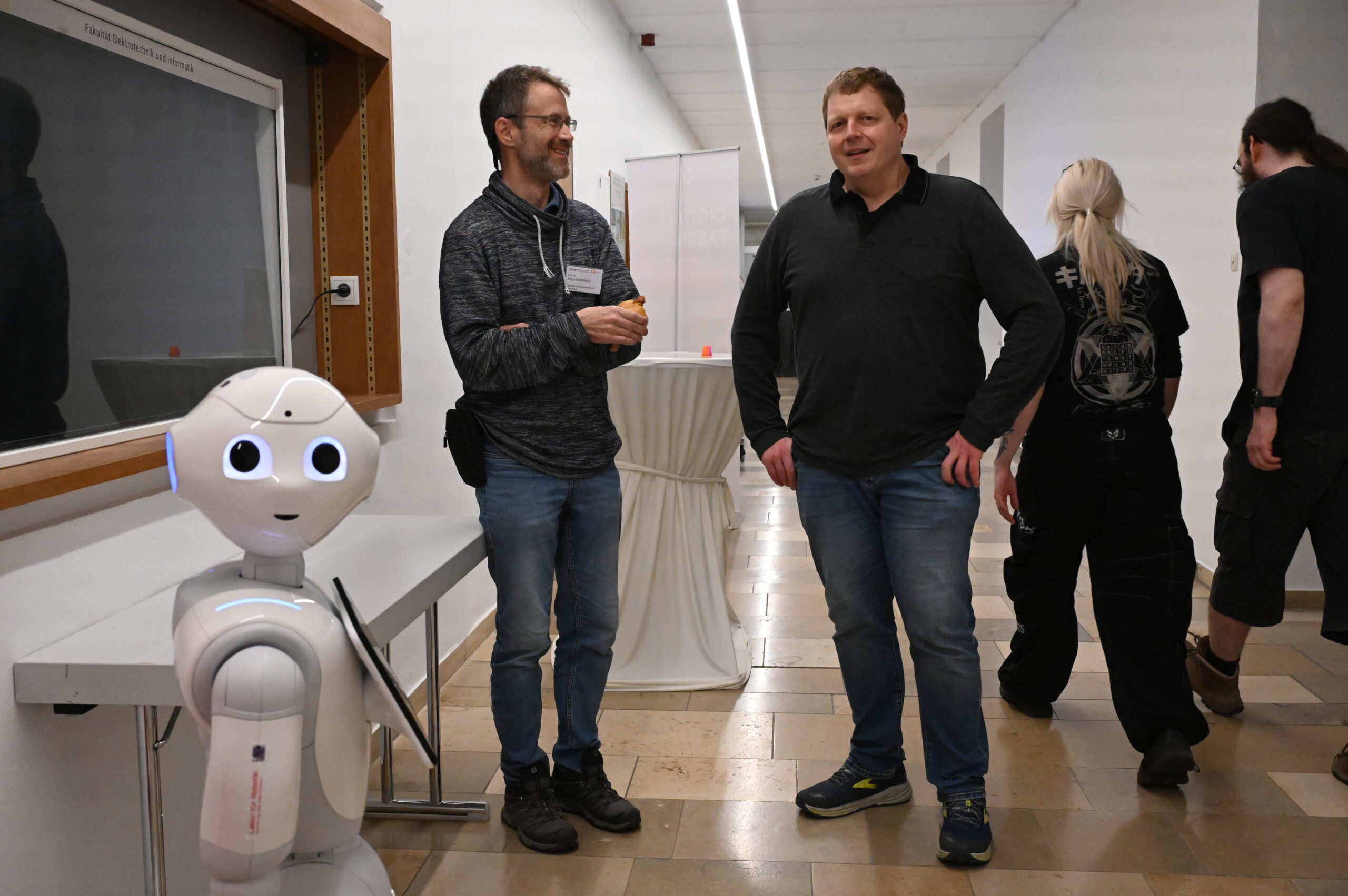 Zwei Männer stehen während der Langen Nacht der Wissenschaften in einem Korridor neben einem weißen humanoiden Roboter und unterhalten sich. Im Hintergrund gehen eine Frau mit langen blonden Haaren und eine weitere Person von dieser Wissenschaftsveranstaltung weg. at Hochschule Coburg
