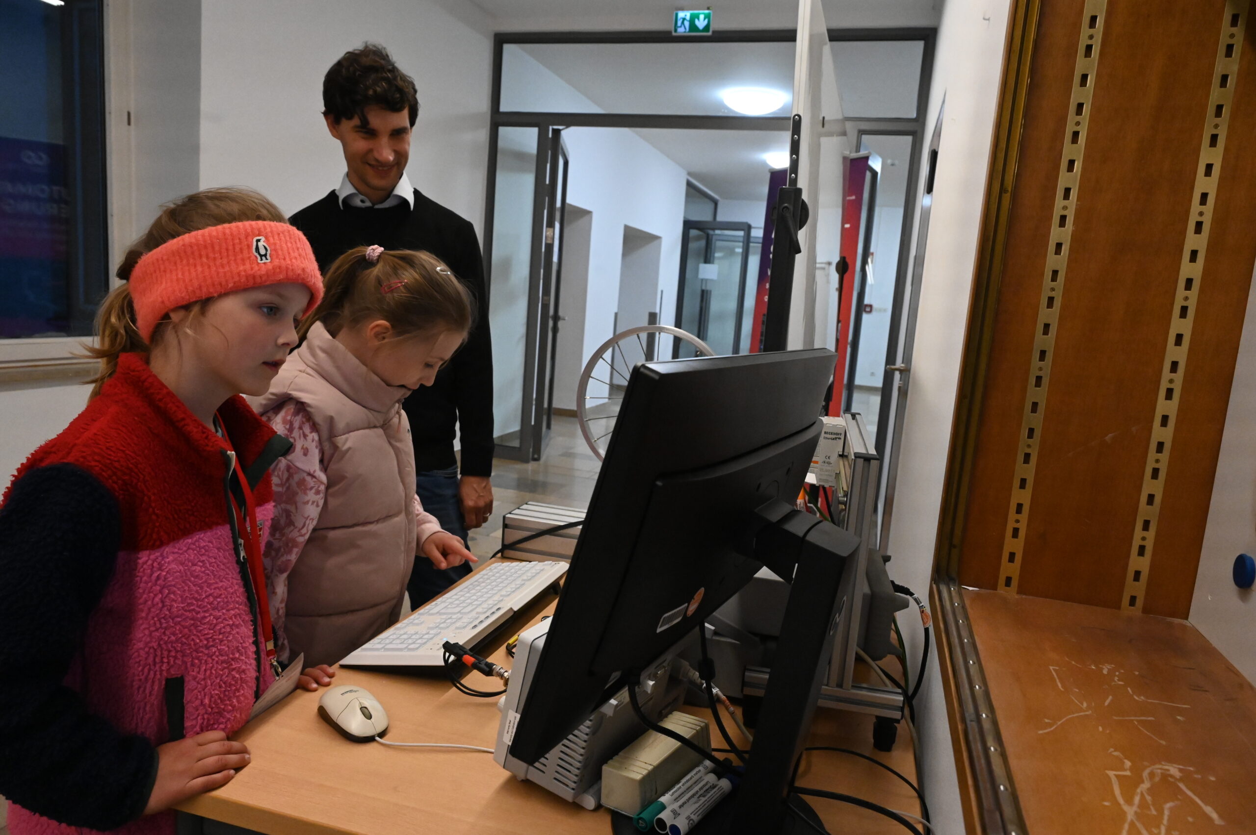 Zwei junge Mädchen benutzen einen Computer, während ein Erwachsener lächelnd hinter ihnen steht. Sie befinden sich in einem Innenraum, in der Nähe eines hölzernen Schreibtisches mit Computerausrüstung, vielleicht erkunden sie Resonanz während der Langen Nacht der Wissenschaften in einem hell erleuchteten Flurbereich. at Hochschule Coburg