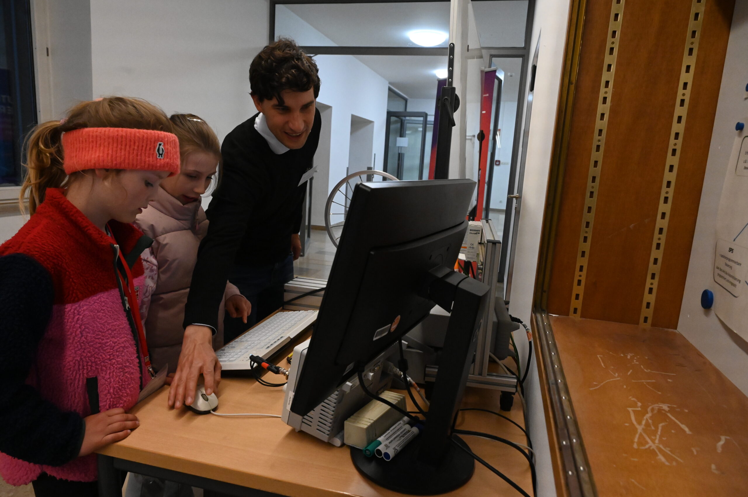 Ein Mann hilft zwei jungen Mädchen während der Langen Nacht der Wissenschaften in einem hell erleuchteten Raum an einem Schreibtisch bei der Benutzung eines Computers. Ein Mädchen trägt ein rotes Stirnband und eine bunte Jacke, das andere einen rosa Mantel. Der Mann zeigt auf den Bildschirm und lächelt. at Hochschule Coburg