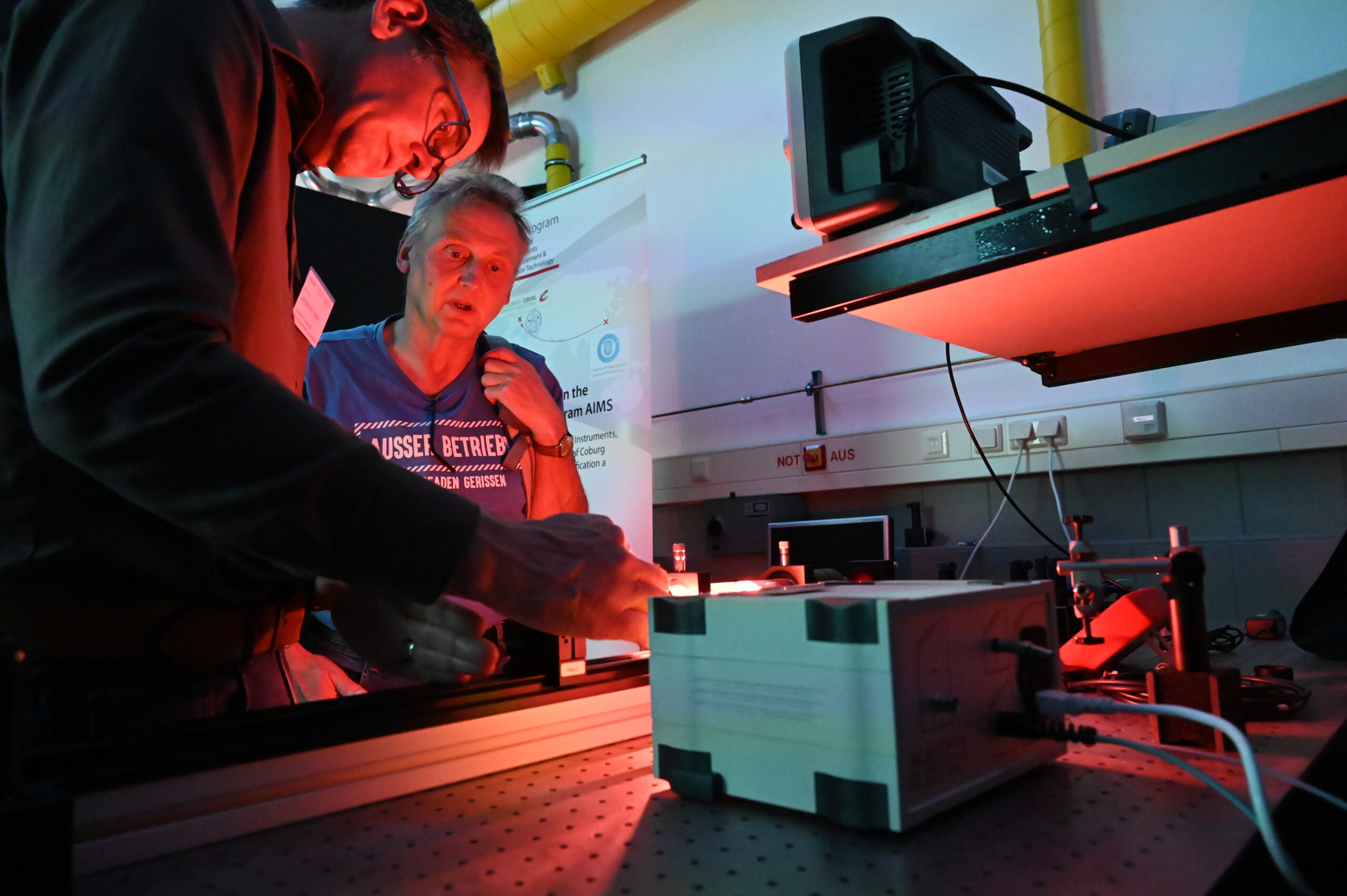 Bei der Langen Nacht der Wissenschaften arbeiten zwei Personen in einem Labor unter roter Beleuchtung mit wissenschaftlichen Geräten. Eine Person hält einen Gegenstand in die Nähe eines Sensors oder einer Kamera, während die andere zuschaut. Auf dem Tisch sind Laborgeräte und Kabel zu sehen. at Hochschule Coburg