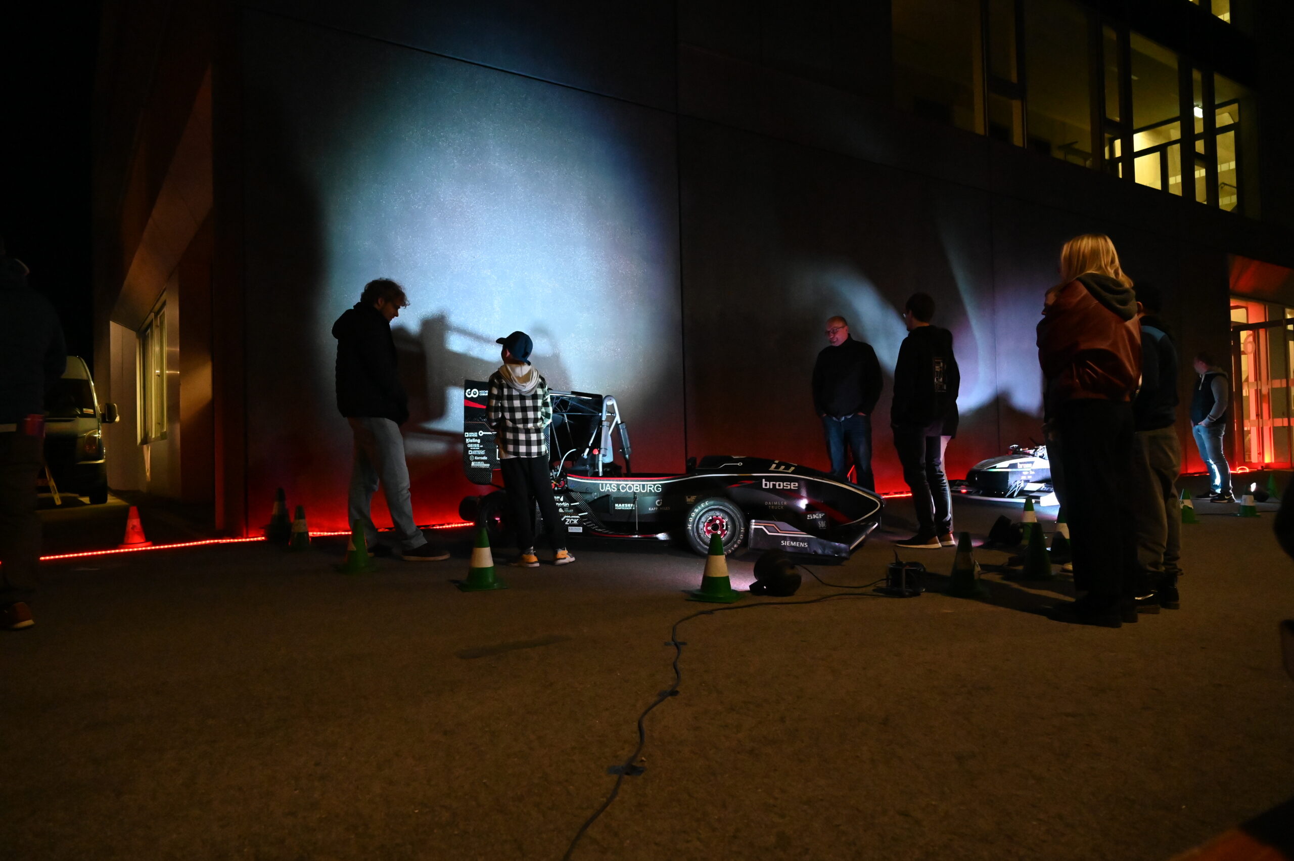 Eine Gruppe von Menschen steht während der Langen Nacht der Wissenschaften auf einer schwach beleuchteten Straße um ein kleines schwarzes Rennauto herum, wobei Schatten auf eine Hauswand geworfen werden und Kegel den Bereich markieren. at Hochschule Coburg
