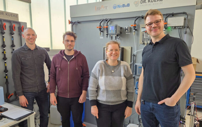 Vier Personen stehen lächelnd vor einer technischen Einrichtung mit Schalttafeln und Rohren, auf denen Logos für LSAT, NoBuB und Dr. Kueke abgebildet sind. Die Gruppe scheint sich in einem Labor oder einem industriellen Arbeitsbereich zu befinden, der auf Forschung und Sensorik ausgerichtet ist. at Hochschule Coburg