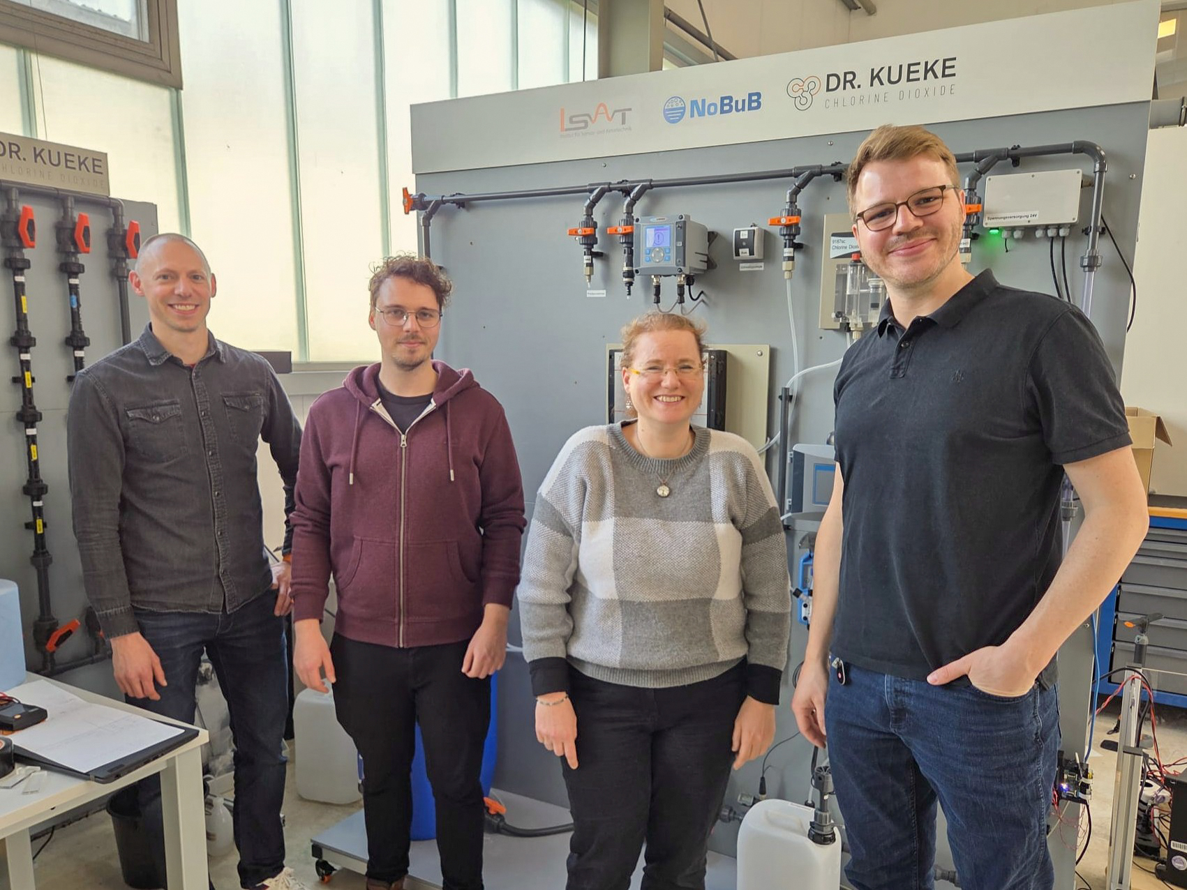 Vier Personen stehen lächelnd vor einer technischen Einrichtung mit Schalttafeln und Rohren, auf denen Logos für LSAT, NoBuB und Dr. Kueke abgebildet sind. Die Gruppe scheint sich in einem Labor oder einem industriellen Arbeitsbereich zu befinden, der auf Forschung und Sensorik ausgerichtet ist. at Hochschule Coburg