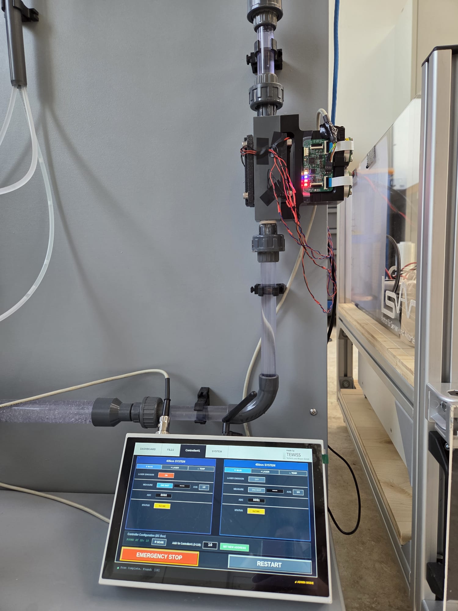 Ein Touchscreen-Bedienfeld zeigt die Systemdaten unterhalb eines vertikalen Rohrs mit angebrachter Sensorik an, die mit einer elektronischen Platine mit LEDs verbunden ist, und verdeutlicht die moderne Wasserdesinfektionstechnologie in einer Labor- oder industriellen Forschungsumgebung. at Hochschule Coburg
