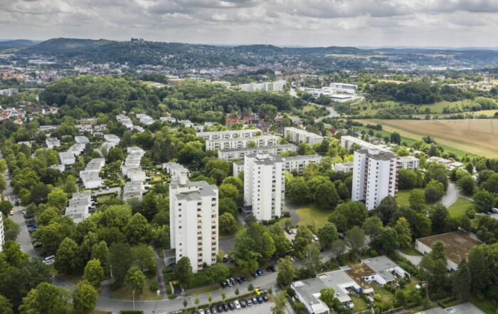Luftaufnahme eines Vorortes mit weißen Wohnhäusern in der Nähe der Hochschule Coburg, umgeben von grünen Bäumen, Feldern und Straßen; der Heimatring schlängelt sich durch die Szene, in der Ferne sind eine Stadt und Hügel unter einem bewölkten Himmel zu sehen. at Hochschule Coburg