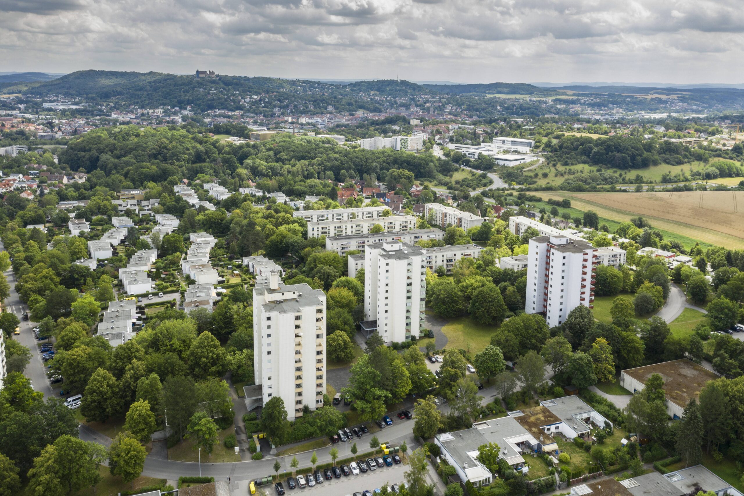 Luftaufnahme eines Vorortes mit weißen Wohnhäusern in der Nähe der Hochschule Coburg, umgeben von grünen Bäumen, Feldern und Straßen; der Heimatring schlängelt sich durch die Szene, in der Ferne sind eine Stadt und Hügel unter einem bewölkten Himmel zu sehen. at Hochschule Coburg
