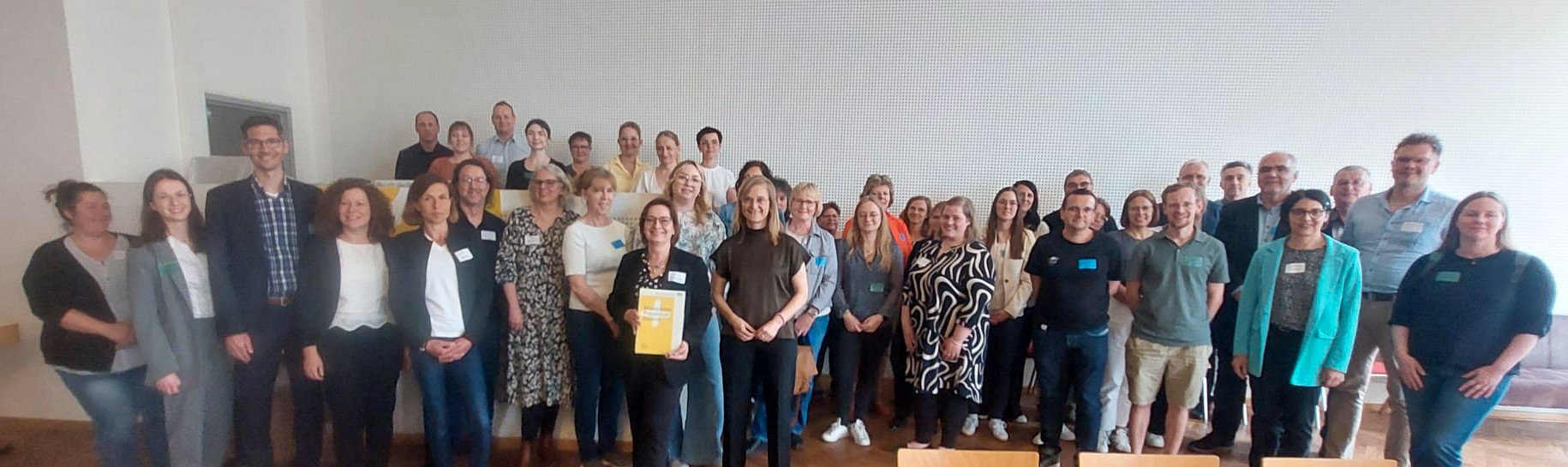 Eine große Gruppe Erwachsener steht in einem Klassenzimmer oder Tagungsraum zusammen und lächelt in die Kamera - einige halten Dokumente zum Thema Prävention in der Hand. Die bunt gemischte Gruppe wirkt engagiert und bereit, mit einer weißen Wand und leeren Stühlen im Hintergrund. at Hochschule Coburg