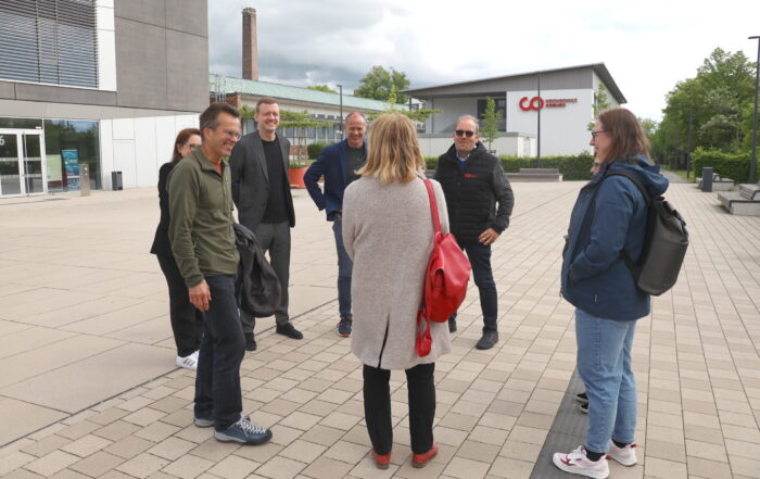 Sieben Erwachsene stehen draußen auf einer gepflasterten Fläche in der Nähe der modernen Gebäude der Hochschule Coburg, unterhalten sich und lächeln. Eine Frau mit einer roten Tasche steht mit dem Rücken zur Kamera, während Grüne Landtagsabgeordnete lässig mit der Gruppe plaudern. at Hochschule Coburg