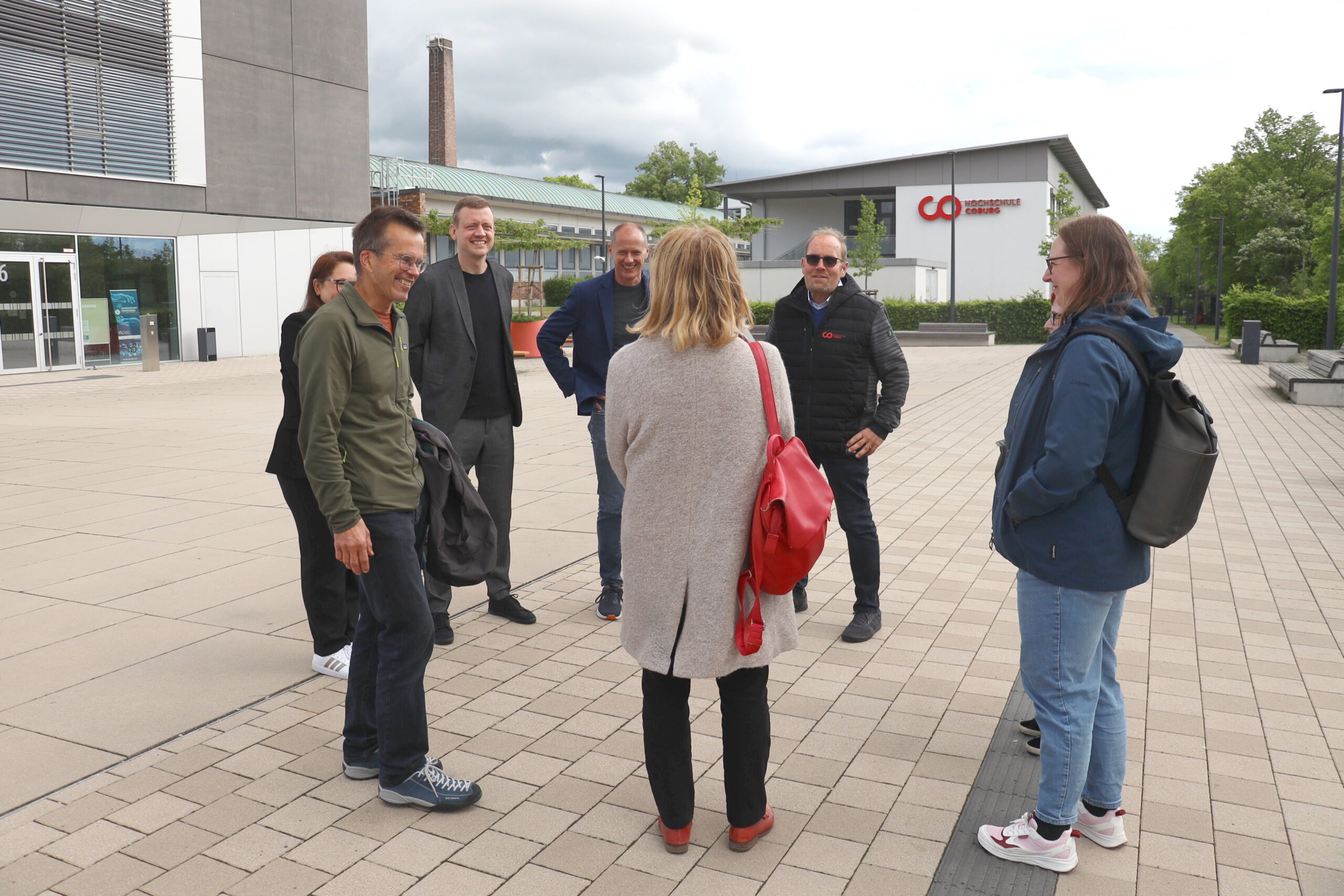 Sieben Erwachsene stehen draußen auf einer gepflasterten Fläche in der Nähe der modernen Gebäude der Hochschule Coburg, unterhalten sich und lächeln. Eine Frau mit einer roten Tasche steht mit dem Rücken zur Kamera, während Grüne Landtagsabgeordnete lässig mit der Gruppe plaudern. at Hochschule Coburg
