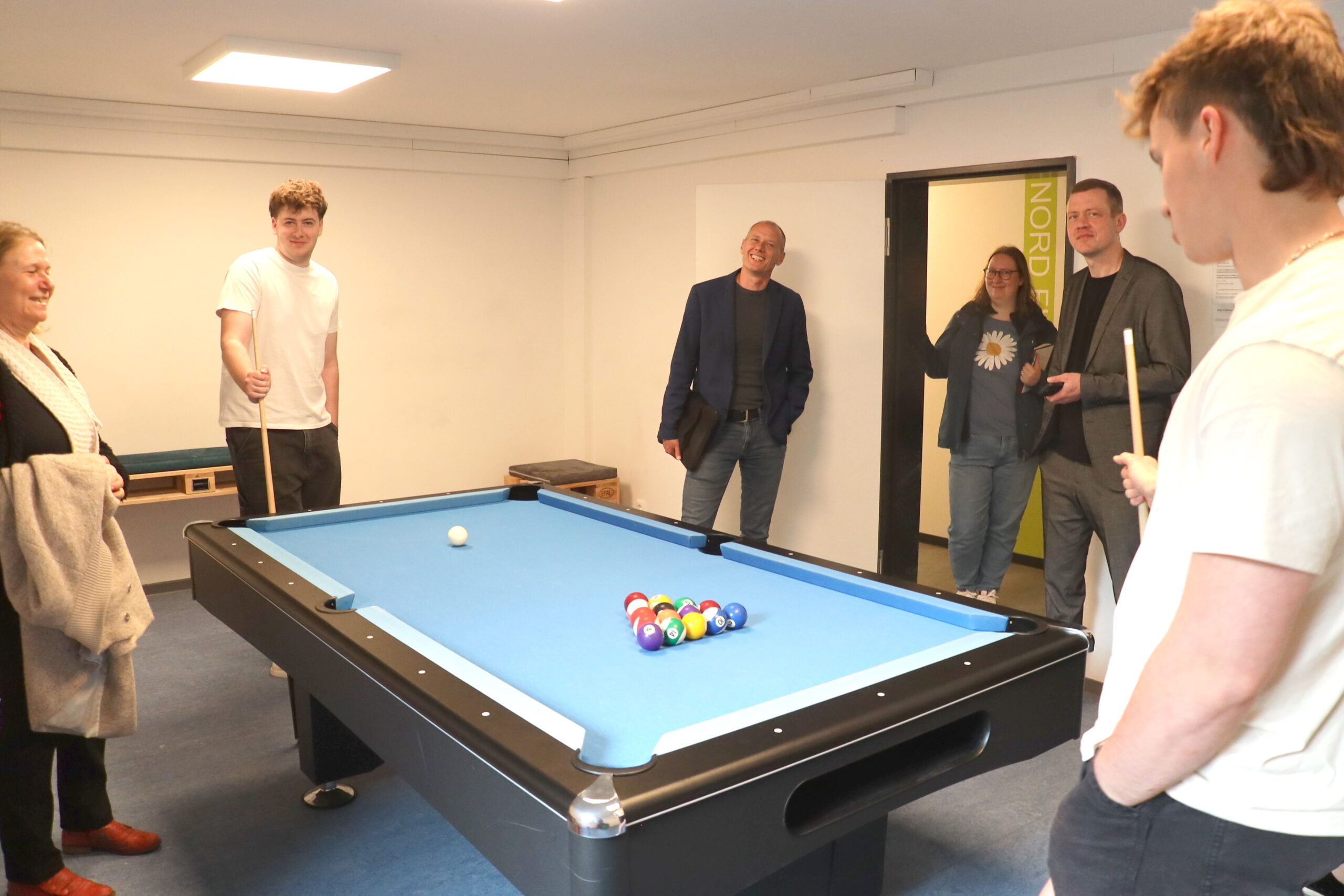 Sechs Personen stehen um einen blauen Billardtisch an der Hochschule Coburg, einige halten Queues in der Hand und bereiten sich in einem gut beleuchteten Raum auf eine Partie Billard vor. Die Atmosphäre wirkt entspannt und freundlich. at Hochschule Coburg