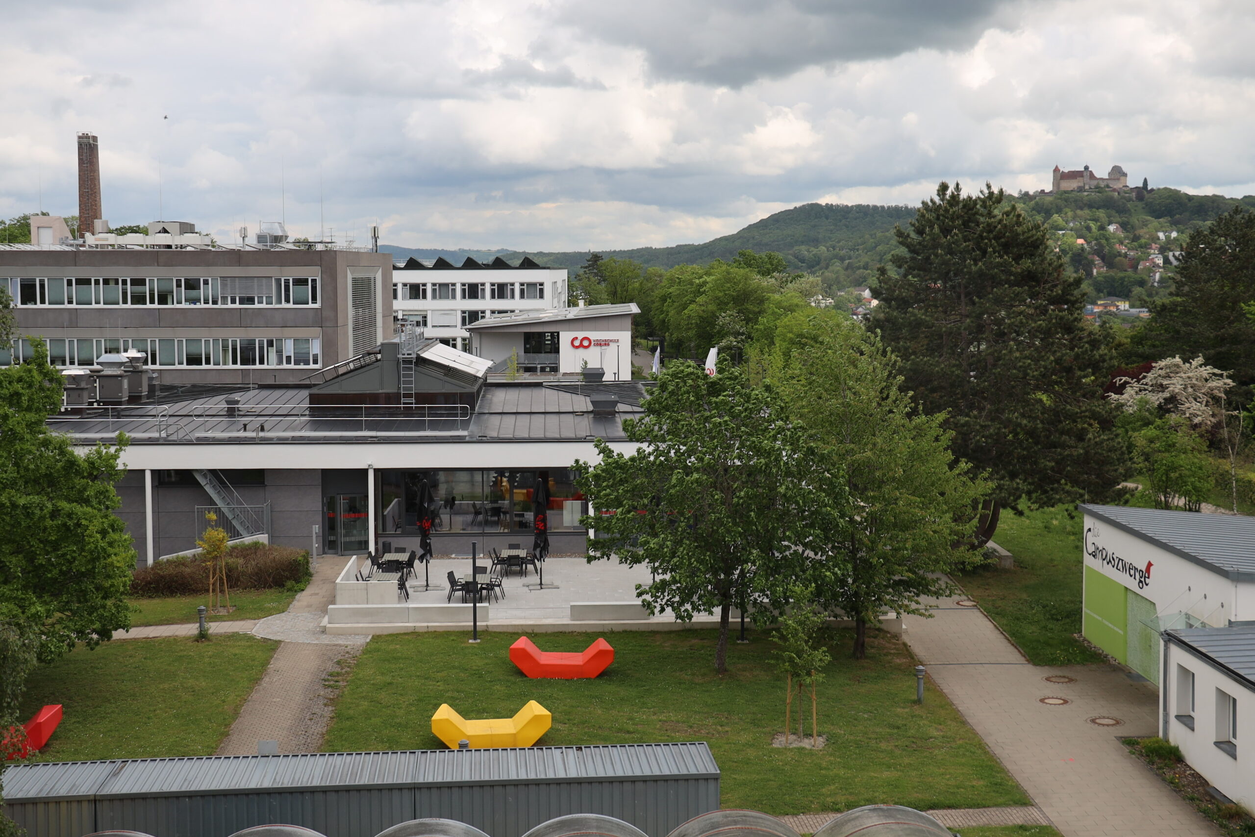 Ein moderner Campus der Hochschule Coburg mit weißen und grauen Gebäuden, Sitzgelegenheiten im Freien, bunten Bänken auf Rasenflächen und grünen Bäumen. Im Hintergrund sind Hügel und ein Schloss bei bewölktem Himmel zu sehen. at Hochschule Coburg