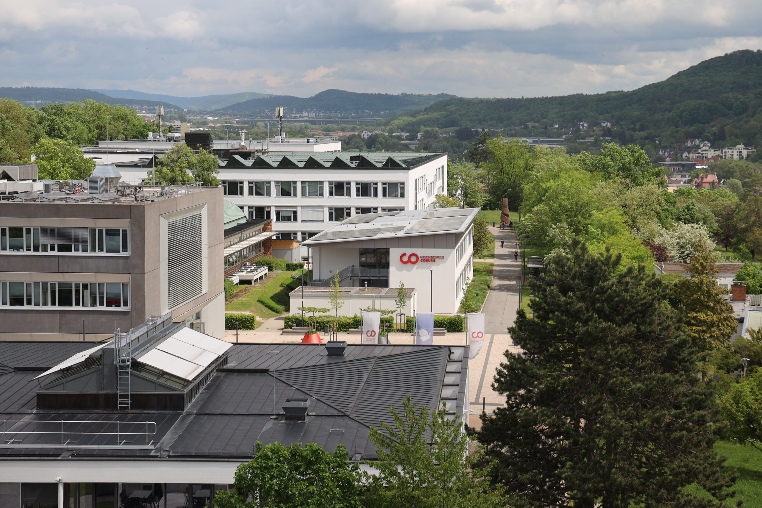 Luftaufnahme des modernen Campus der Hochschule Coburg mit mehreren Gebäuden, grünen Bäumen und Hügeln im Hintergrund unter einem teilweise bewölkten Himmel. In der Mitte ist ein Gebäude mit einem roten "CO"-Logo zu sehen, in dem die Grünen Landtagsabgeordneten arbeiten. at Hochschule Coburg