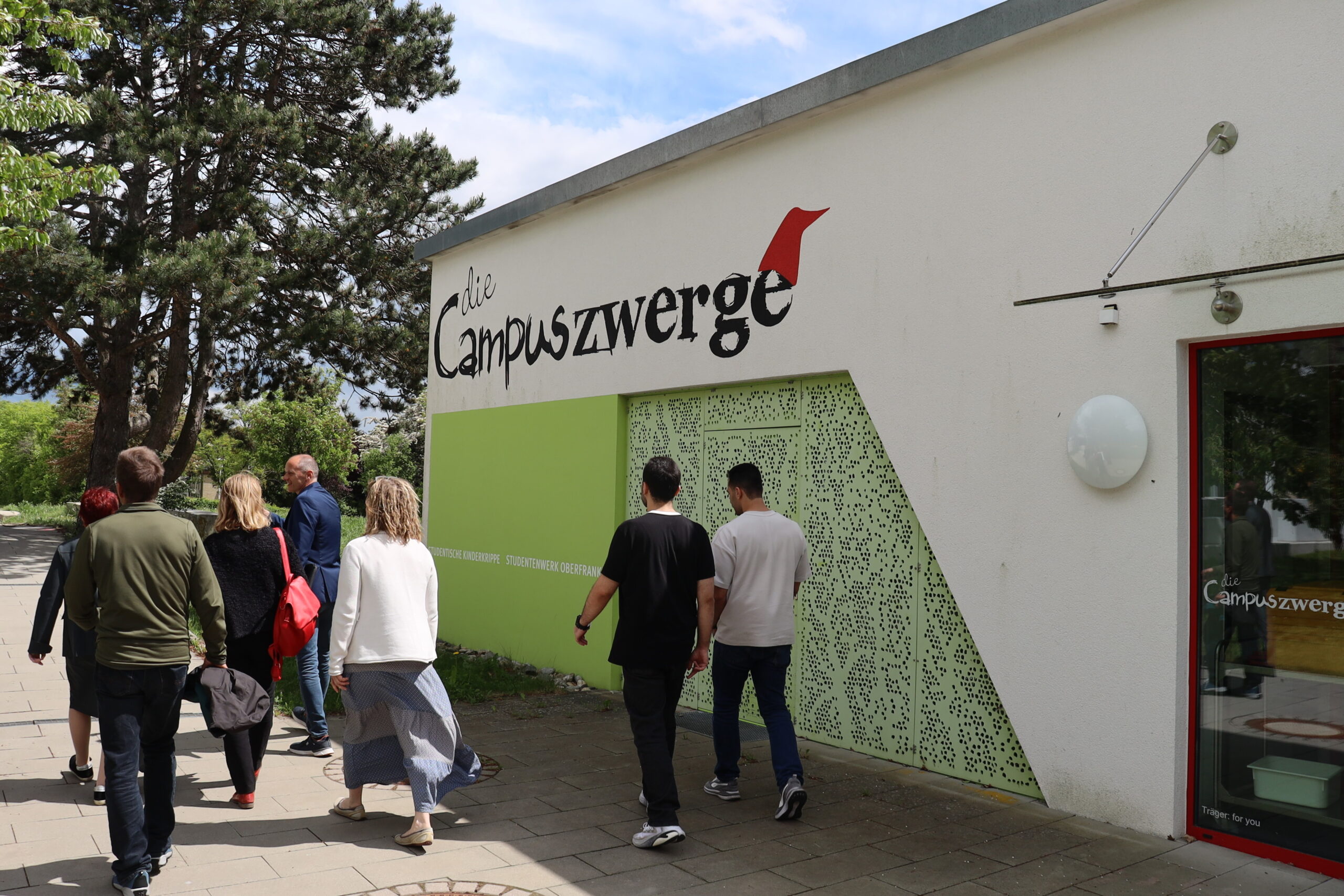 Eine Gruppe von Personen, darunter auch Grüne Landtagsabgeordnete, geht auf ein weißes Gebäude zu, auf dem in großen Buchstaben "die Campuszwerge" steht, daneben ein grüner Abschnitt und eine rot umrahmte Tür. Die Szene spielt im Freien in der Nähe der Hochschule Coburg, mit Bäumen und einem gefliesten Gehweg. at Hochschule Coburg