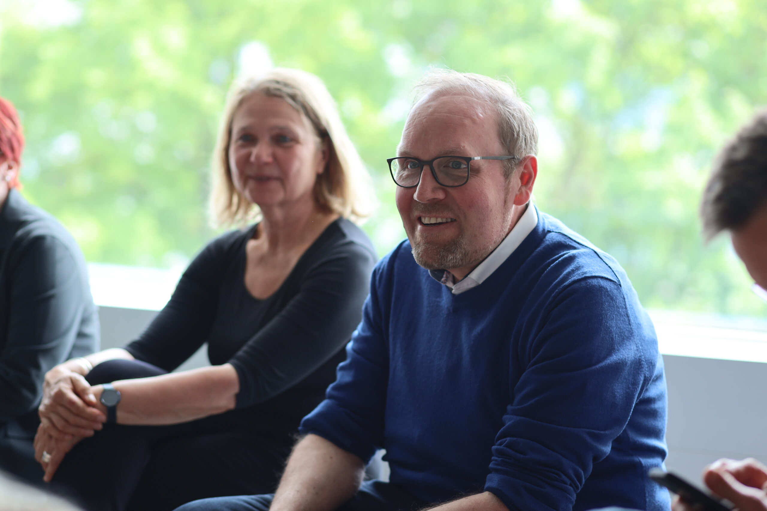Ein Mann mit Brille und blauem Pullover lächelt, während er in einem Haus neben einer Frau in Schwarz sitzt, möglicherweise eine Grüne Landtagsabgeordnete. Durch ein Fenster sind grüne Bäume zu sehen - vielleicht an der Hochschule Coburg. at Hochschule Coburg