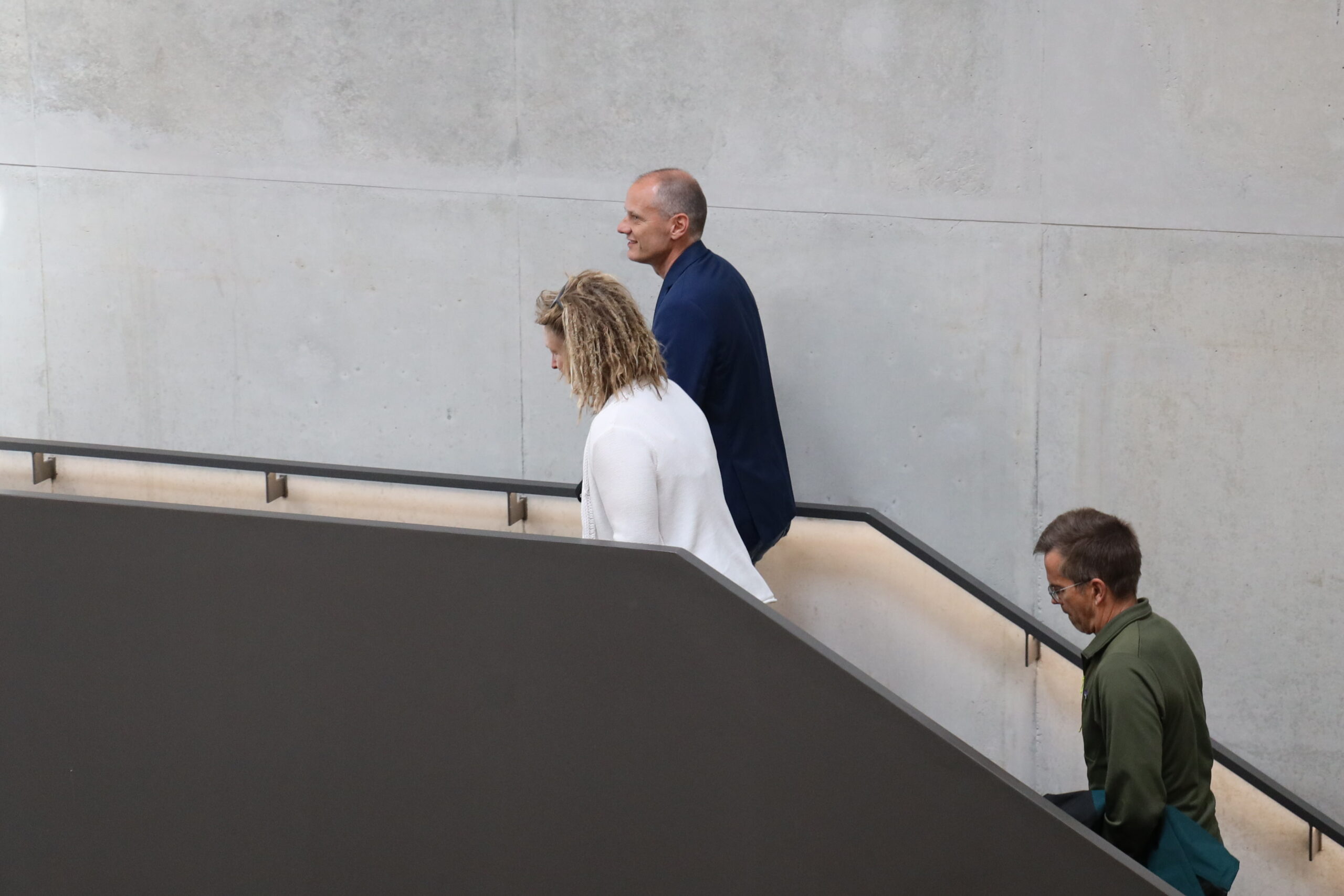 Drei Personen gehen eine Treppe an einer glatten Betonwand der Hochschule Coburg hinauf; eine trägt weiß, eine andere blau, und die dritte, möglicherweise ein grün gekleideter Grüner Landtagsabgeordneter, hält sich am Handlauf fest. at Hochschule Coburg
