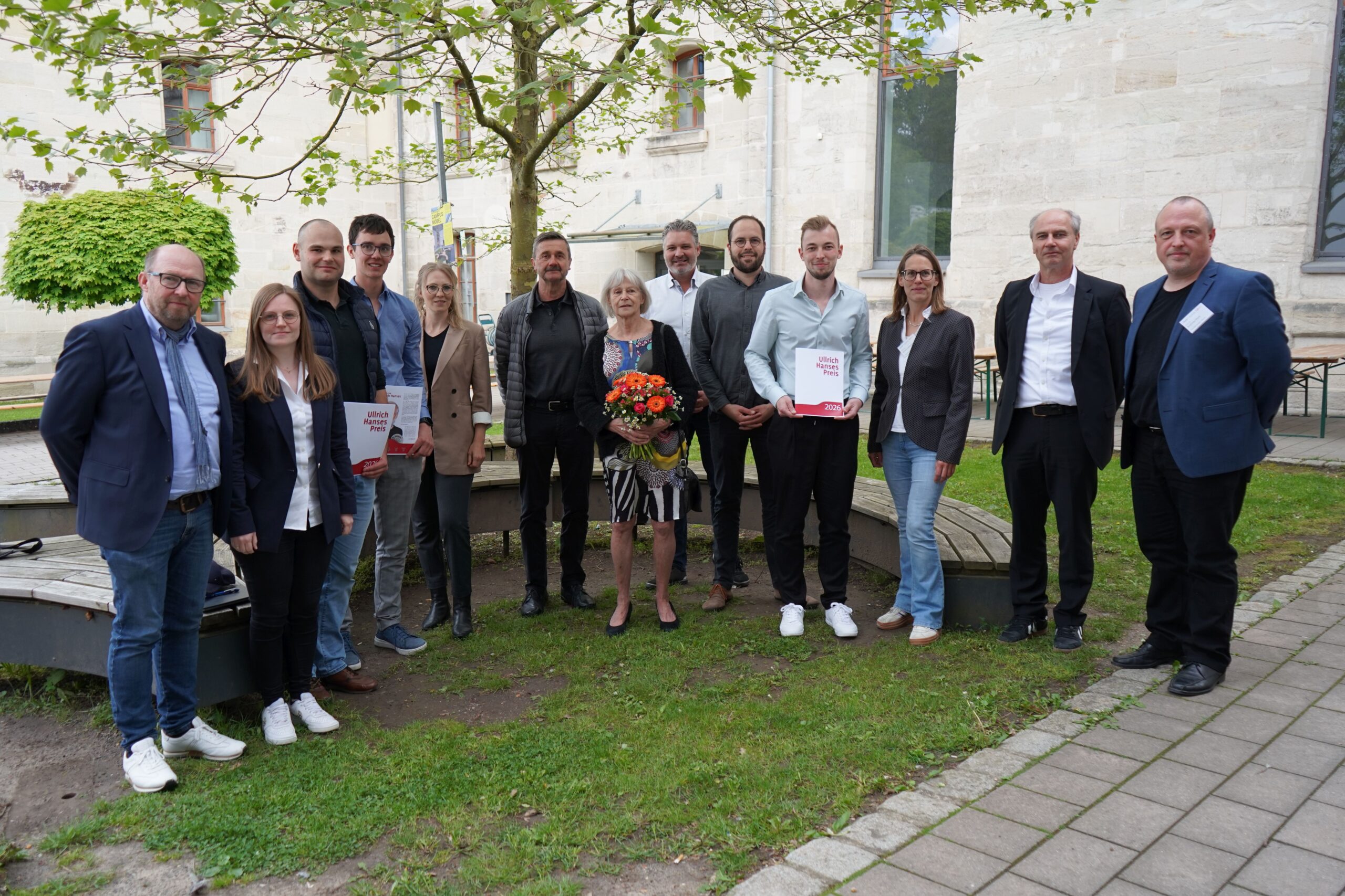 Eine Gruppe von vierzehn Personen steht im Freien vor einem steinernen Gebäude. Die Frau in der Mitte hält einen Blumenstrauß in der Hand, während mehrere Personen Urkunden für Spitzenleistungen im Bauingenieurwesen zeigen, die möglicherweise mit dem Kurbasik- und Hanses-Preis ausgezeichnet wurden. at Hochschule Coburg