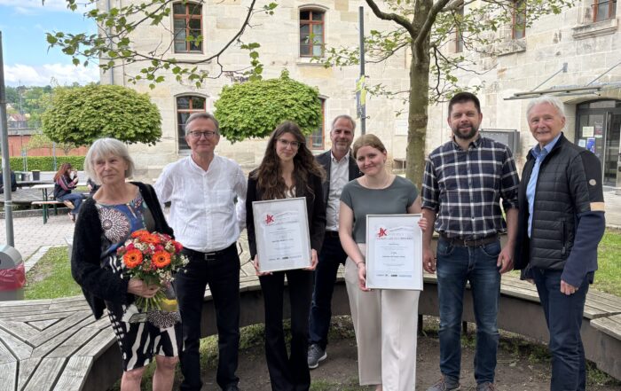 Sieben Personen posieren im Freien; zwei Frauen in der Mitte halten gerahmte Kurbasik- und Hanses-Preis-Urkunden, die Spitzenleistungen im Bauingenieurwesen auszeichnen. Eine Frau auf der linken Seite hält einen Blumenstrauß, im Hintergrund ein altes Steingebäude und Bäume. at Hochschule Coburg
