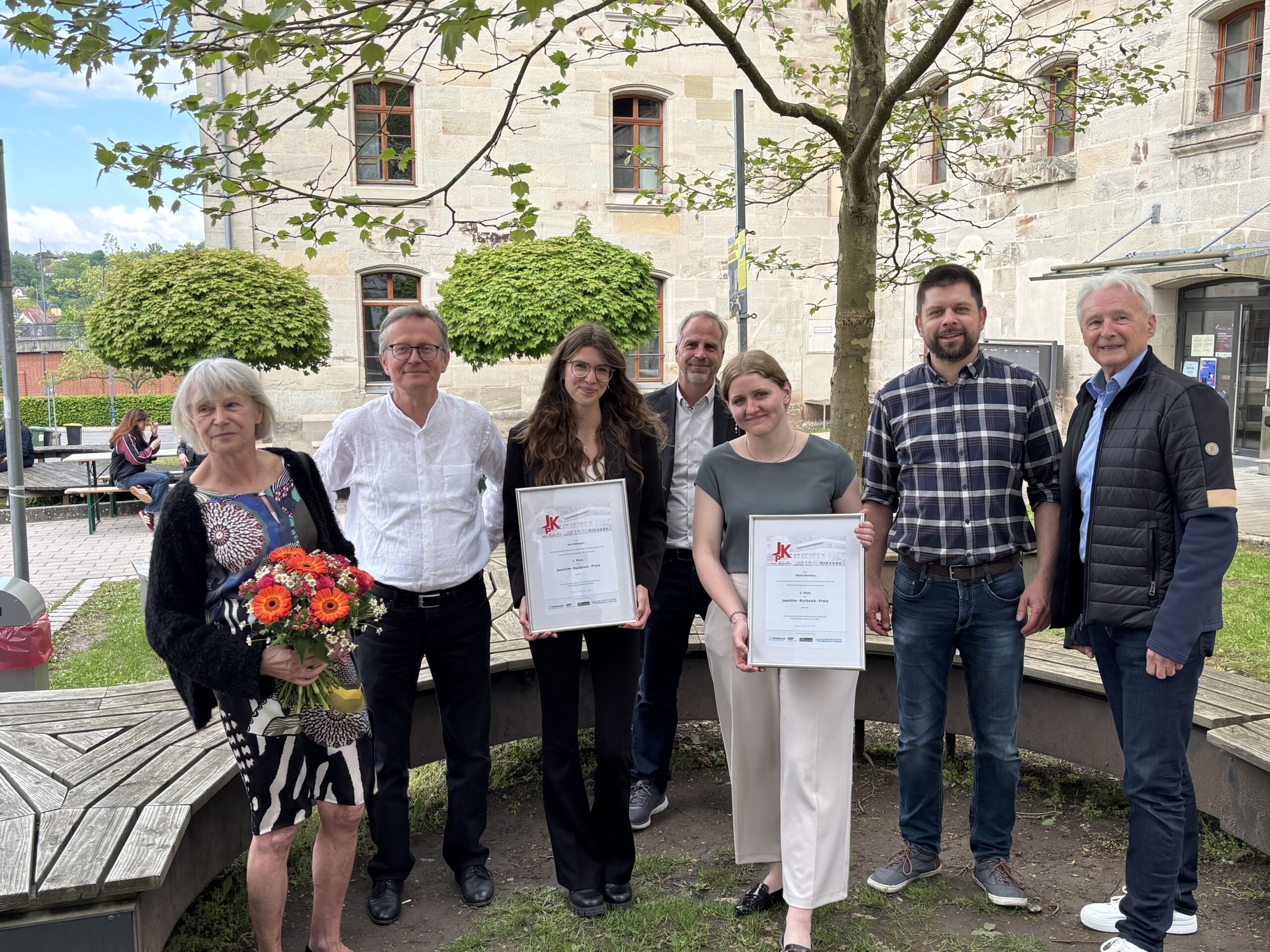 Sieben Personen posieren im Freien; zwei Frauen in der Mitte halten gerahmte Kurbasik- und Hanses-Preis-Urkunden, die Spitzenleistungen im Bauingenieurwesen auszeichnen. Eine Frau auf der linken Seite hält einen Blumenstrauß, im Hintergrund ein altes Steingebäude und Bäume. at Hochschule Coburg